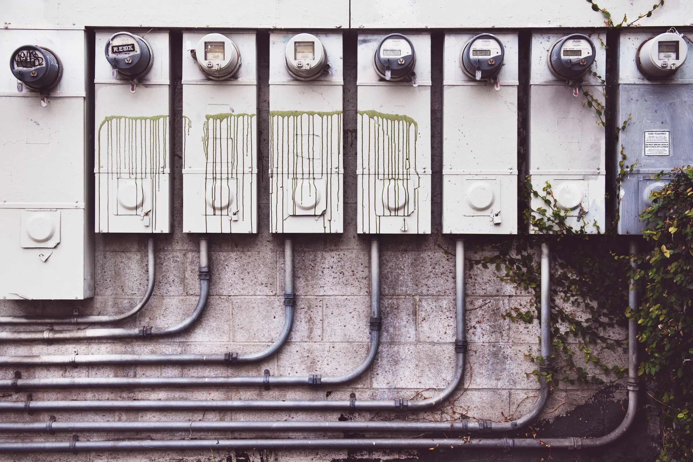 A row of residential electric meters mounted on an exterior wall with metal conduit running down to the ground