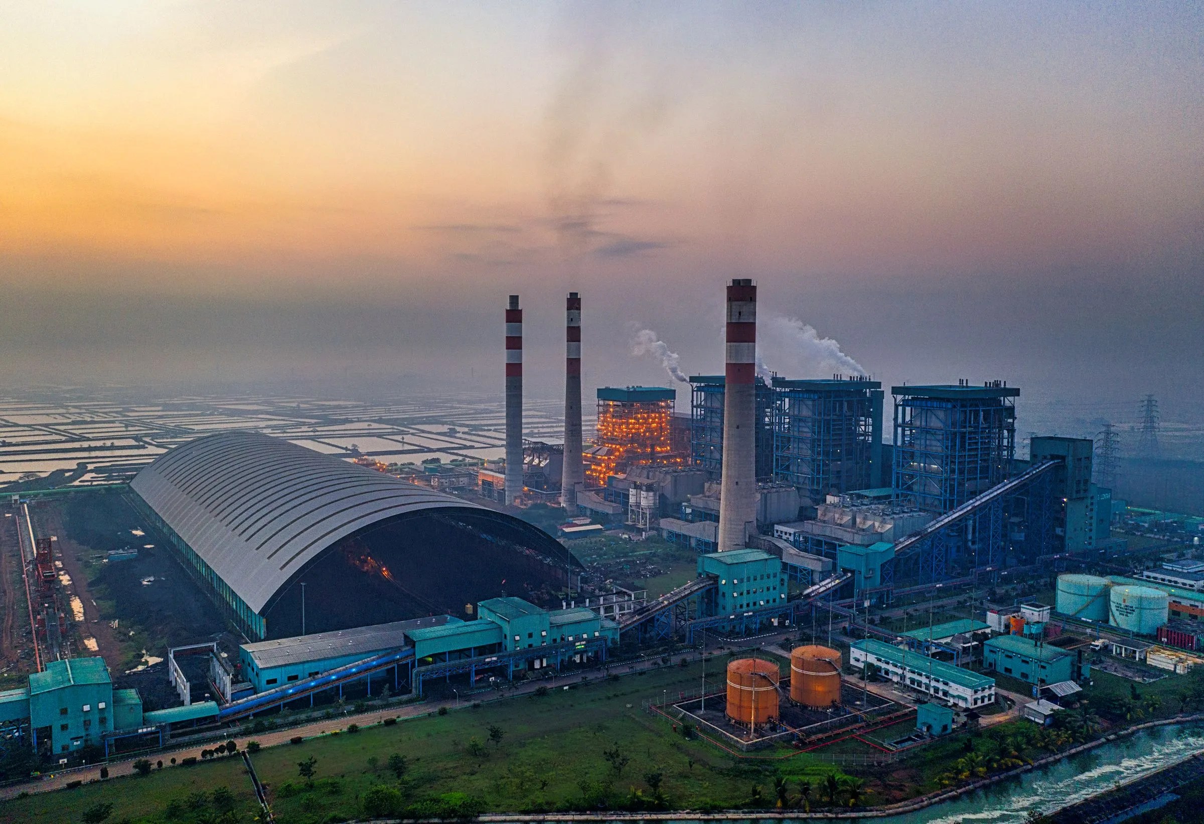 Aerial view of a large thermal power station at dusk, with tall red-and-white smokestacks, boiler buildings, and a domed fuel storage hall