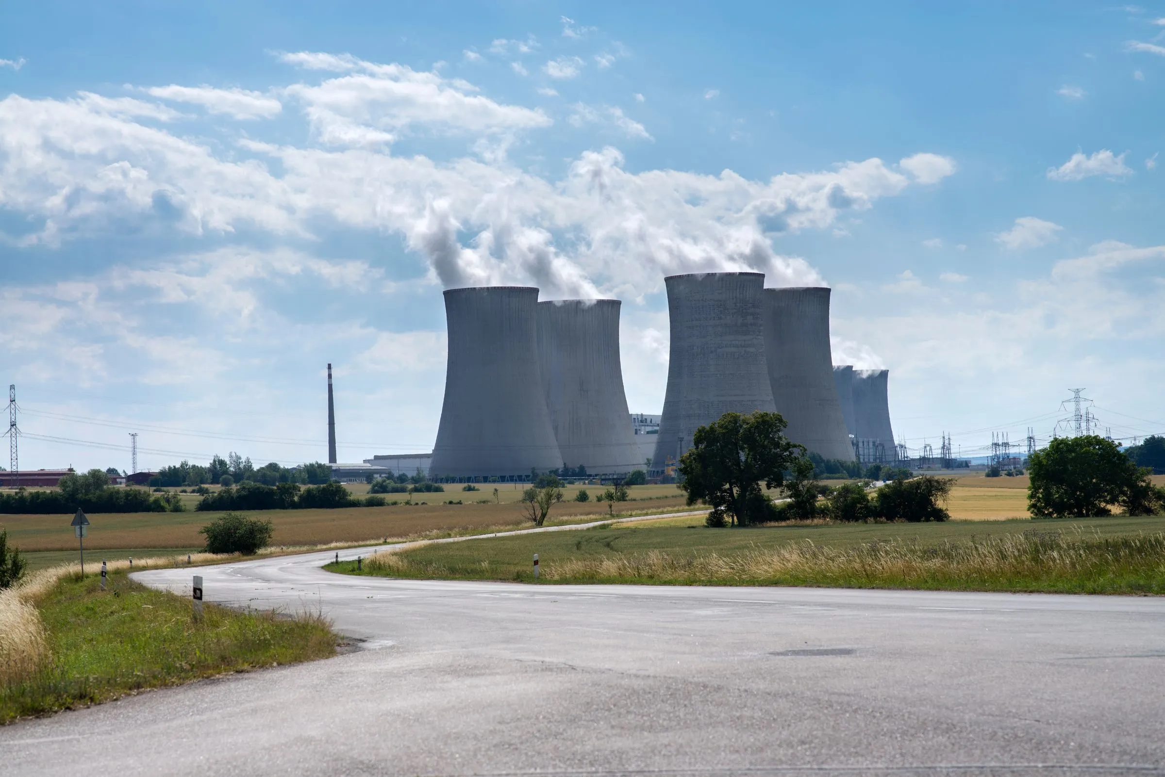 Concrete cooling towers of a nuclear power plant releasing steam into a blue sky, seen from a country road across open fields