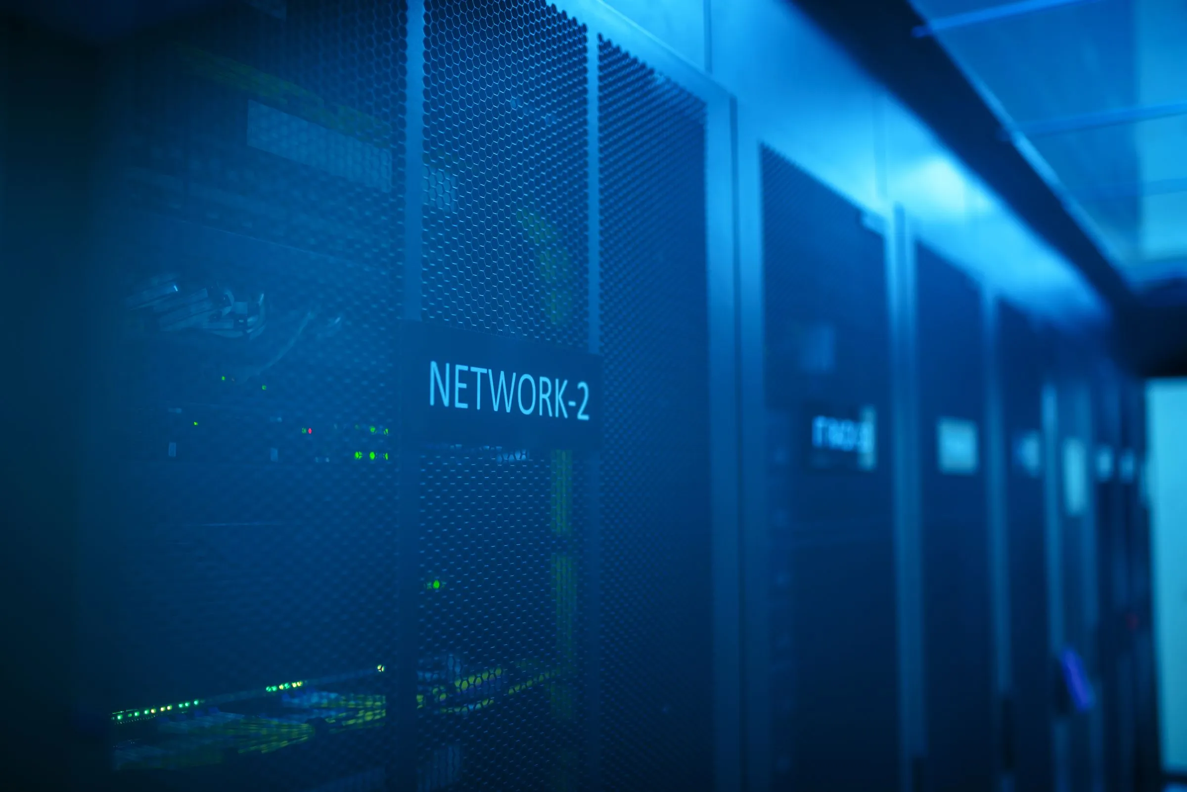 A dark server room bathed in deep blue light, with a row of network cabinets receding into the distance and green status LEDs glowing behind their mesh doors