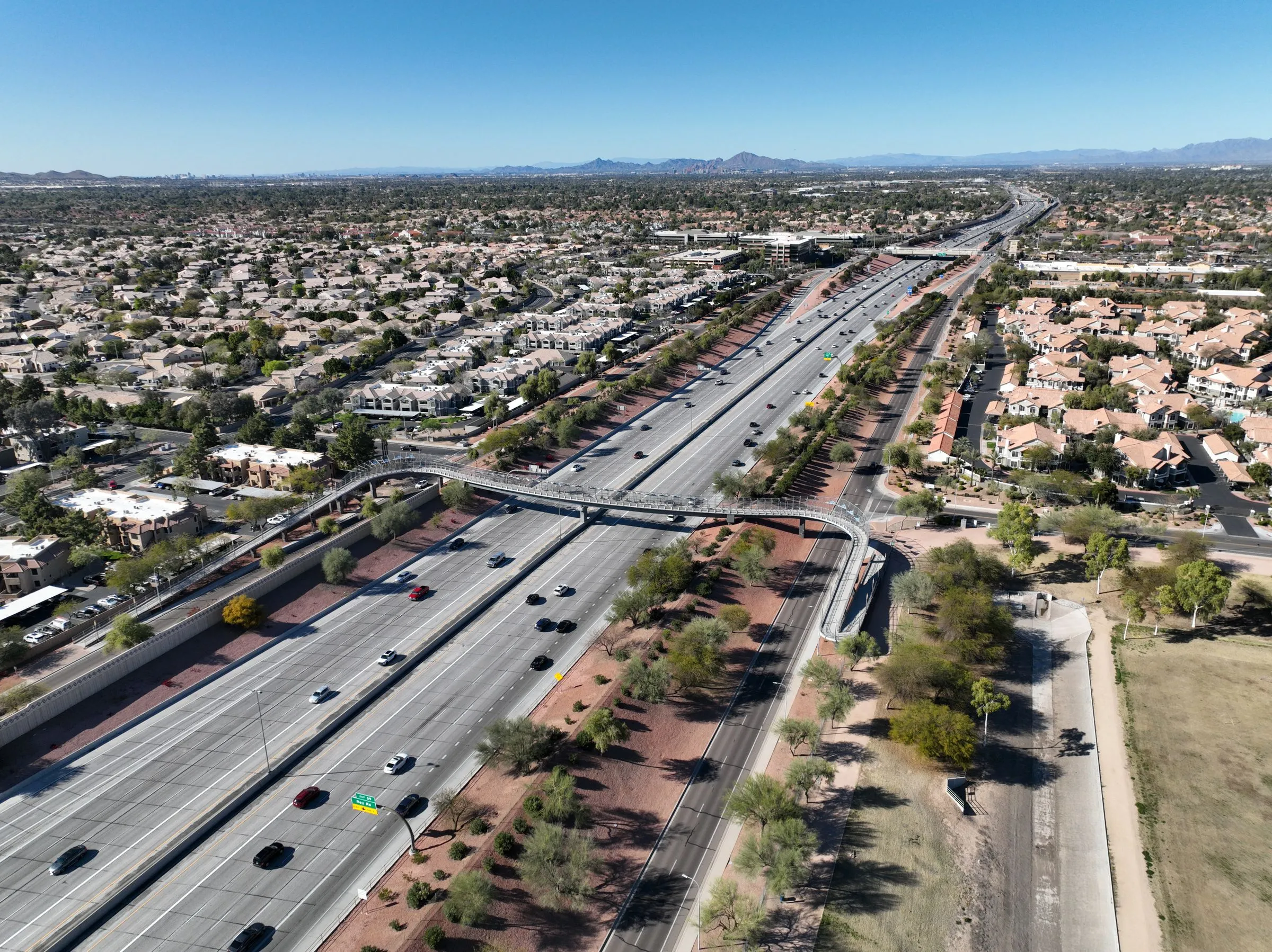 Aerial view of the Loop 101 freeway running through Chandler, Arizona, with a pedestrian bridge crossing the lanes, tile-roofed subdivisions on both sides, and desert mountains on the horizon