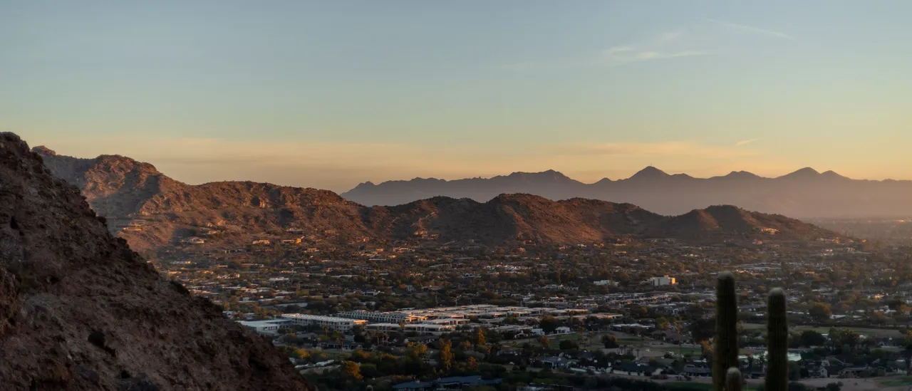 The Phoenix metro area seen from a rocky slope of Camelback Mountain at sunset, with saguaro cacti in the foreground, desert mountains catching the last light, and the city grid stretching to the horizon