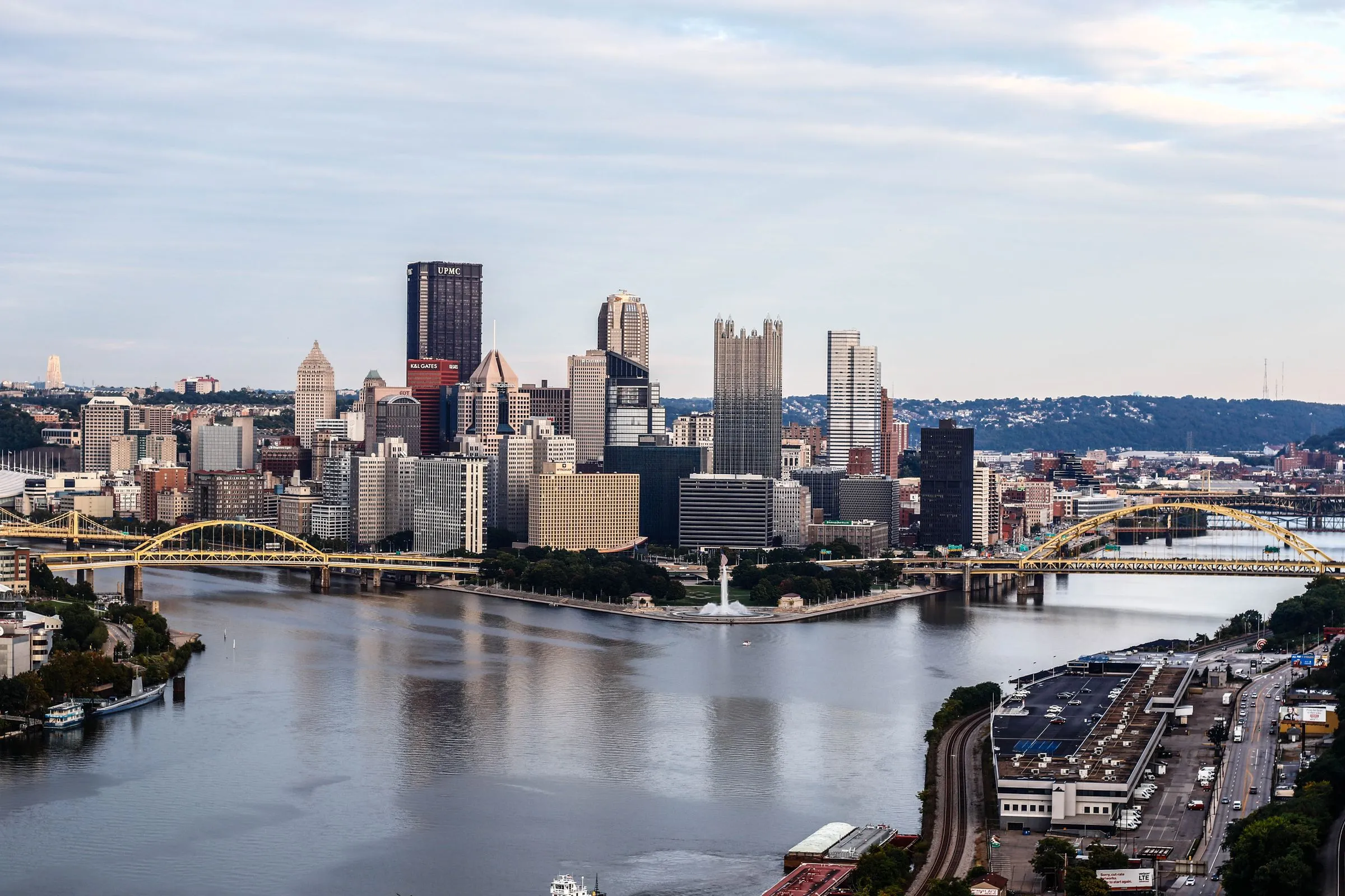 The downtown Pittsburgh skyline from the west on an overcast day, with the Allegheny River at left and the Monongahela at right meeting at the fountain of Point State Park, two yellow steel bridges crossing the rivers, glass and stone office towers behind them, and wooded hills in the distance
