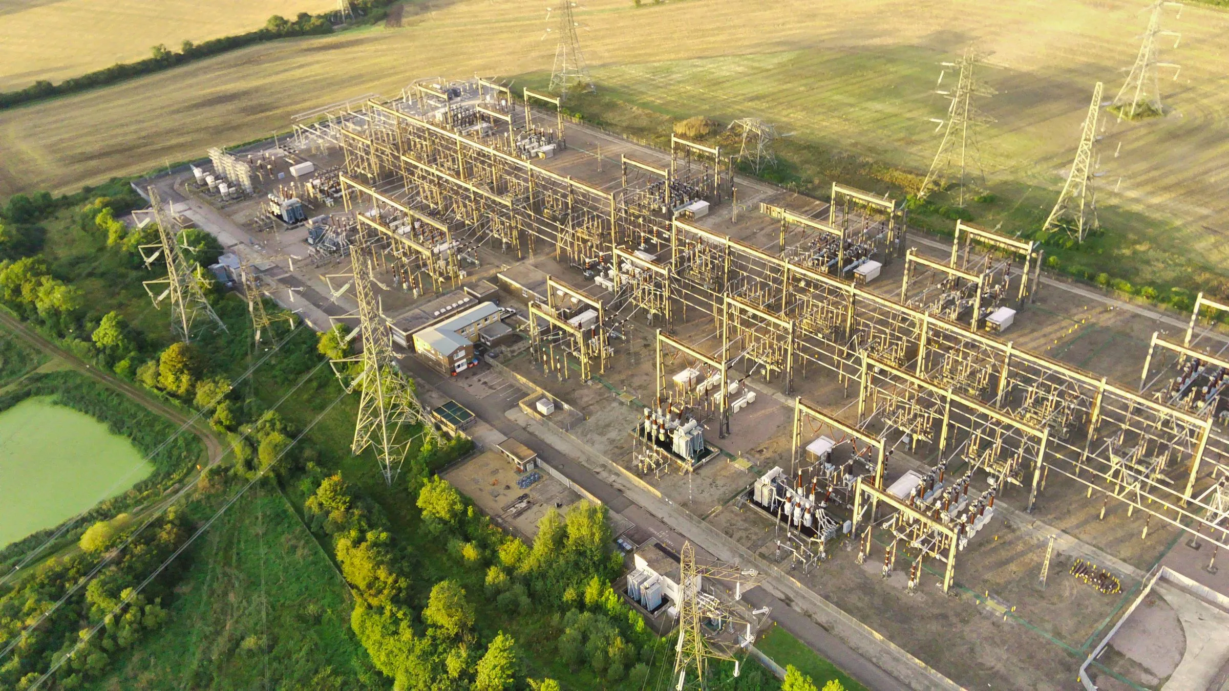 Aerial view of a large high-voltage substation set among harvested farm fields in late-day light, with rows of steel gantries, breakers and transformers inside a fenced yard, lattice transmission towers carrying lines out across the fields, a small control building, and trees and a pond along one edge