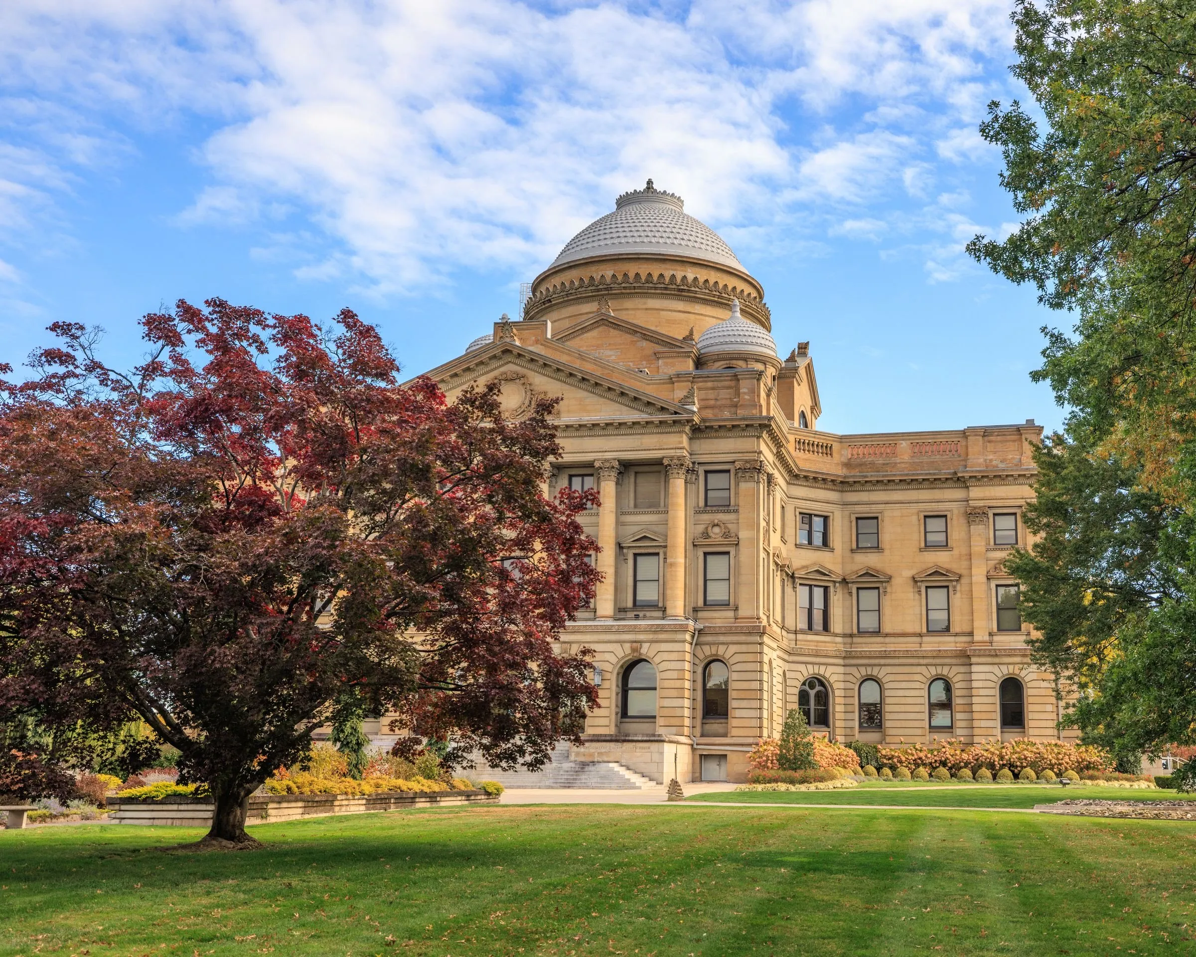 The Luzerne County Courthouse in Wilkes-Barre, Pennsylvania, a Beaux-Arts stone building with a domed central tower and smaller domes, seen across a lawn in autumn with a red-leaved maple in the foreground and a blue sky with scattered clouds