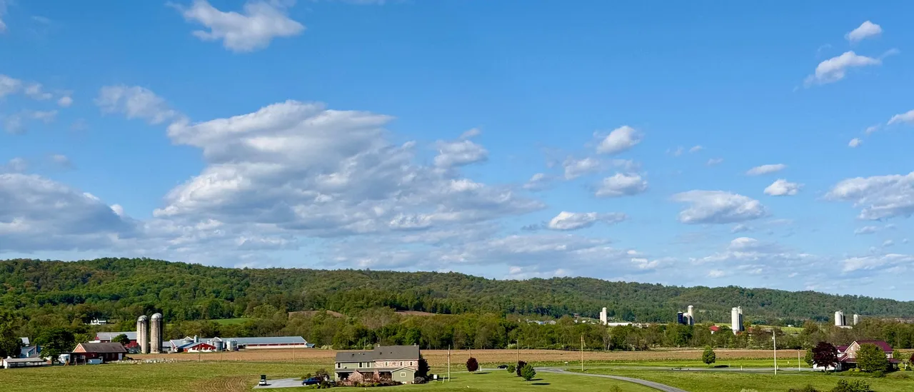 Pennsylvania farm country on a spring afternoon, with a freshly turned field in the foreground, a white farmhouse and red-roofed barns with concrete silos in the middle distance, a two-lane road, and a long forested Appalachian ridge running across the horizon under a blue sky full of cumulus clouds
