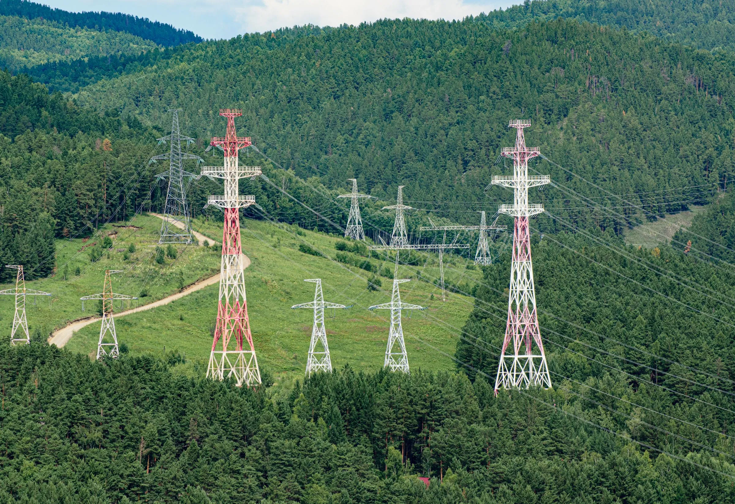 High-voltage transmission towers carrying power lines through forested hills, the kind of grid infrastructure new projects wait years to connect to