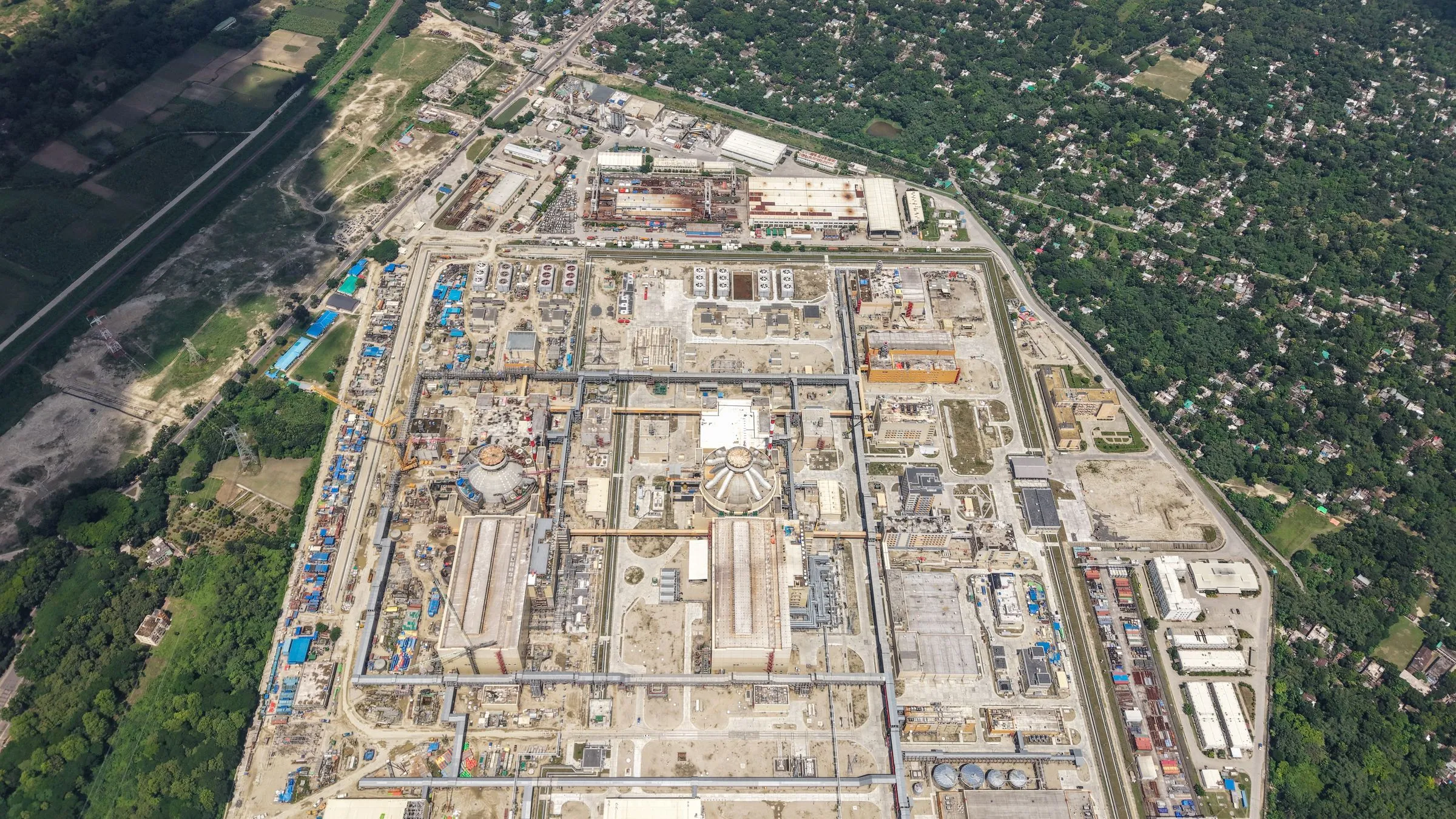 Aerial view of a nuclear power plant construction site with two reactor containment domes rising among cranes, laydown yards, and surrounding greenery