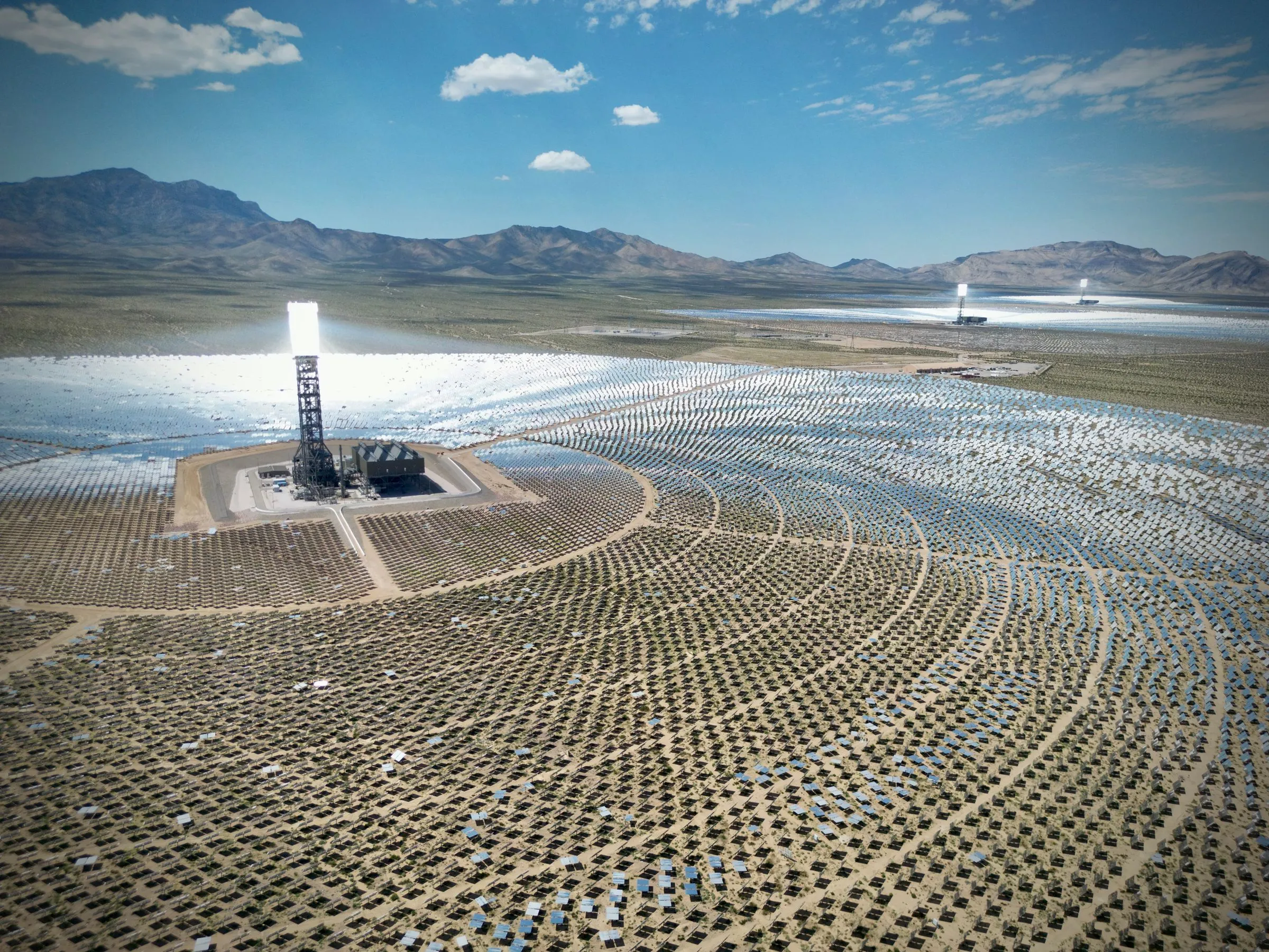 Aerial view of the Ivanpah Solar Electric Generating System in California’s Mojave Desert, with a bright central receiver tower rising from concentric rings of mirrored heliostats on tan desert scrub, two more towers in the distance and bare mountains under a blue sky