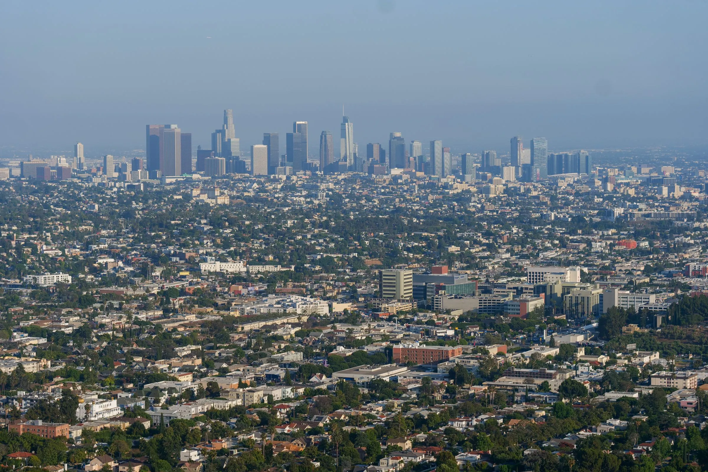 Aerial view of the downtown Los Angeles skyline under a hazy blue sky, with the cluster of high-rise towers rising above miles of low-rise neighborhoods, apartment blocks and trees in the foreground