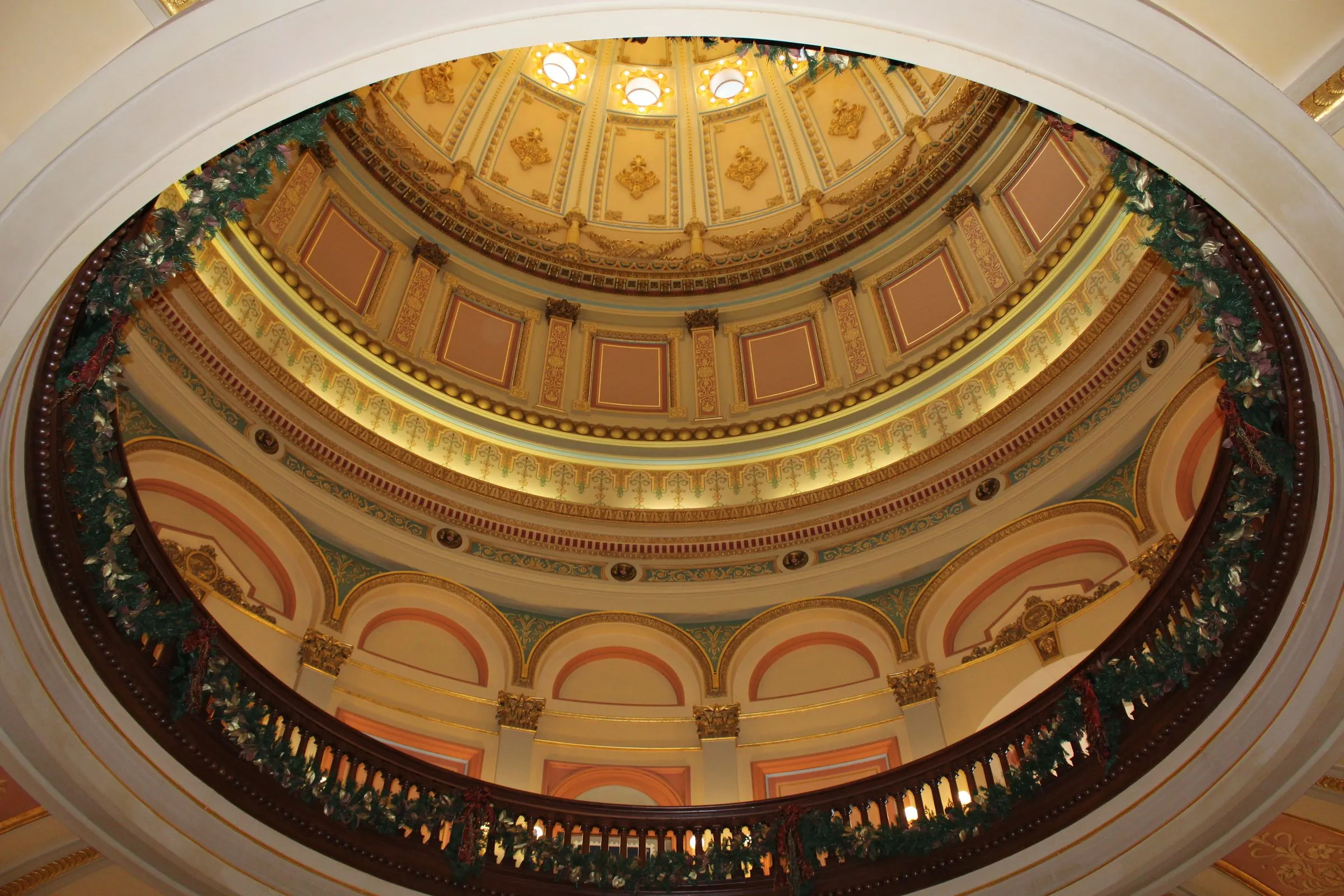 Looking up into the ornate interior of the California State Capitol rotunda in Sacramento, with tiers of gilded arches, painted panels and Corinthian columns rising to the coffered underside of the dome, and a garland-draped railing in the foreground