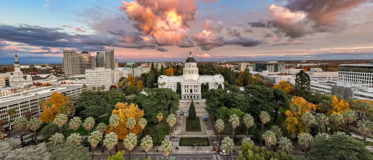 Aerial view of the white neoclassical California State Capitol and its dome in Sacramento, framed by the palms and autumn-yellow trees of Capitol Park with downtown office towers behind under a pink and grey sunset sky