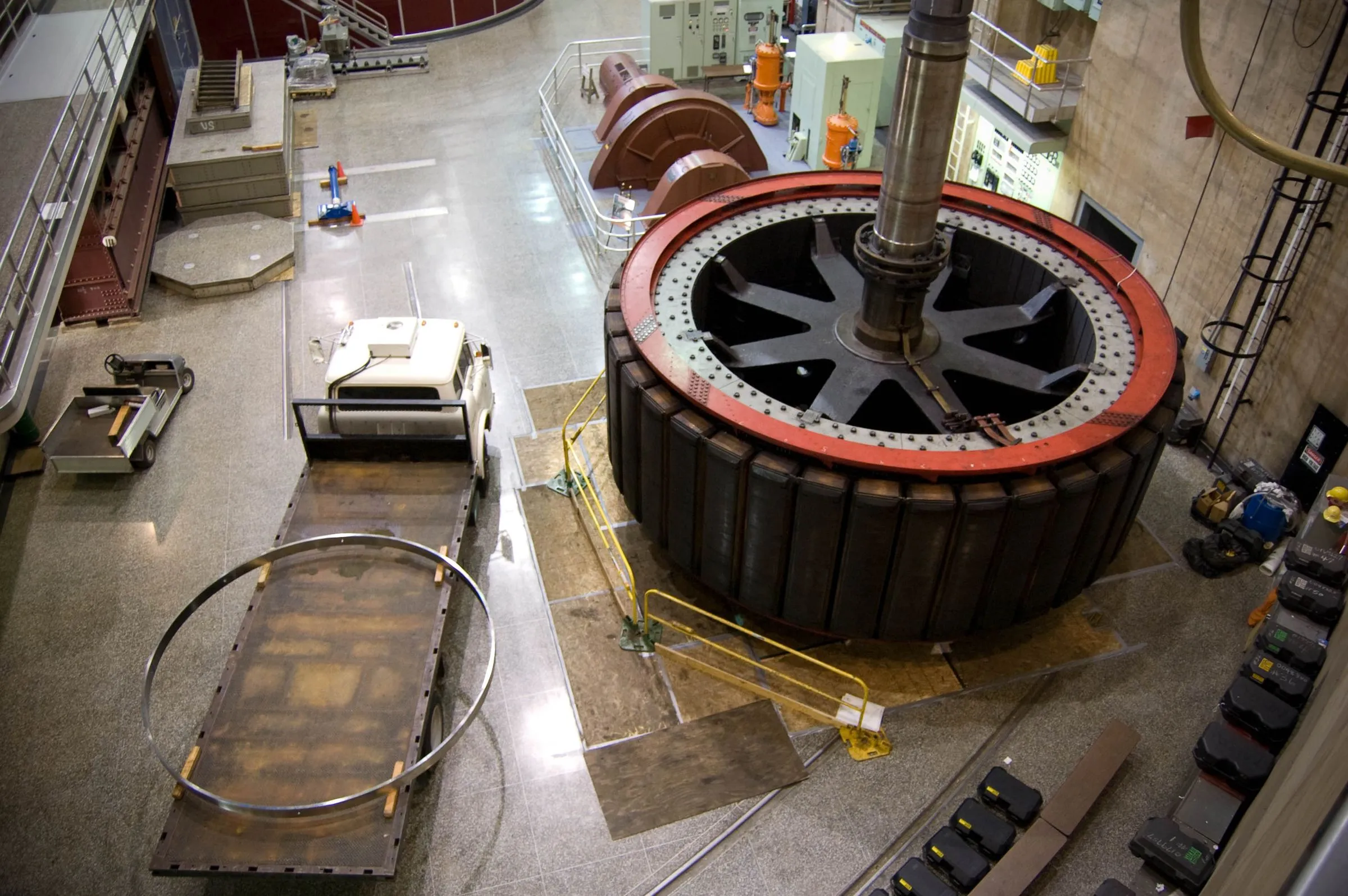 High-angle view of a powerhouse floor with a large generator rotor, a red-rimmed steel ring several meters across with dark laminated poles around its circumference and a vertical shaft rising from its hub, beside a flatbed cart carrying a steel hoop and a row of control cabinets along the far wall