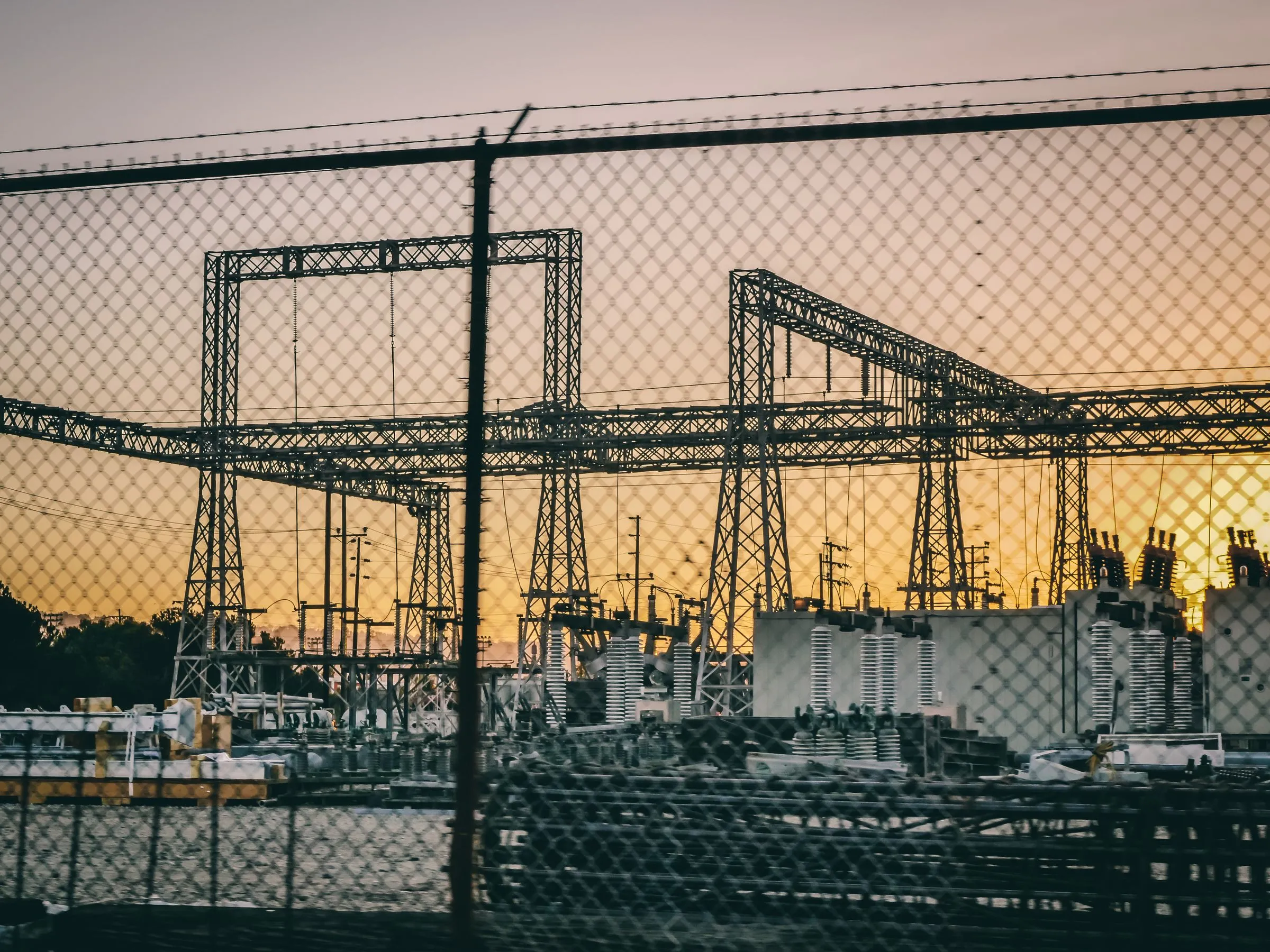A high-voltage switchyard seen through a chain-link fence at dusk, with lattice steel gantries carrying bus conductors, rows of porcelain insulators on transformers in the right foreground, wooden utility poles behind, and an orange sky over trees