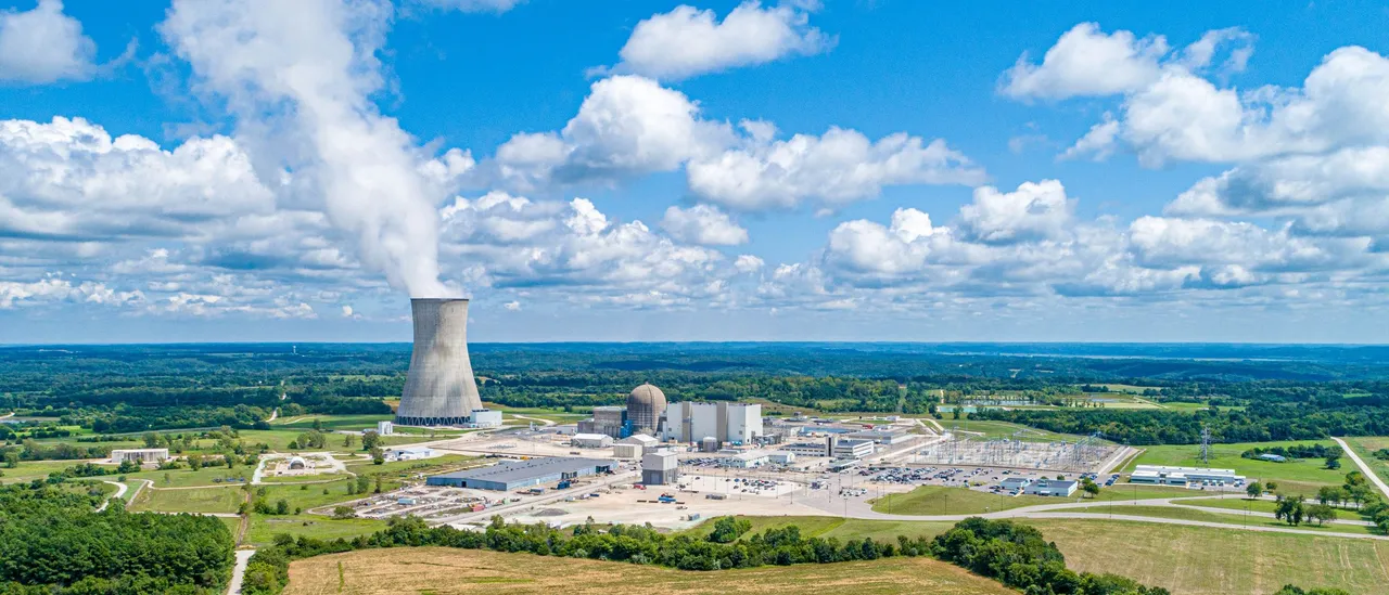 Aerial view of a nuclear power plant on flat Midwestern farmland, with a single concrete cooling tower on the left releasing a white steam plume, a domed concrete containment building and boxy turbine building in the center, parking lots, a switchyard on the right, a plowed field and treeline in the foreground, and cumulus clouds in a blue sky