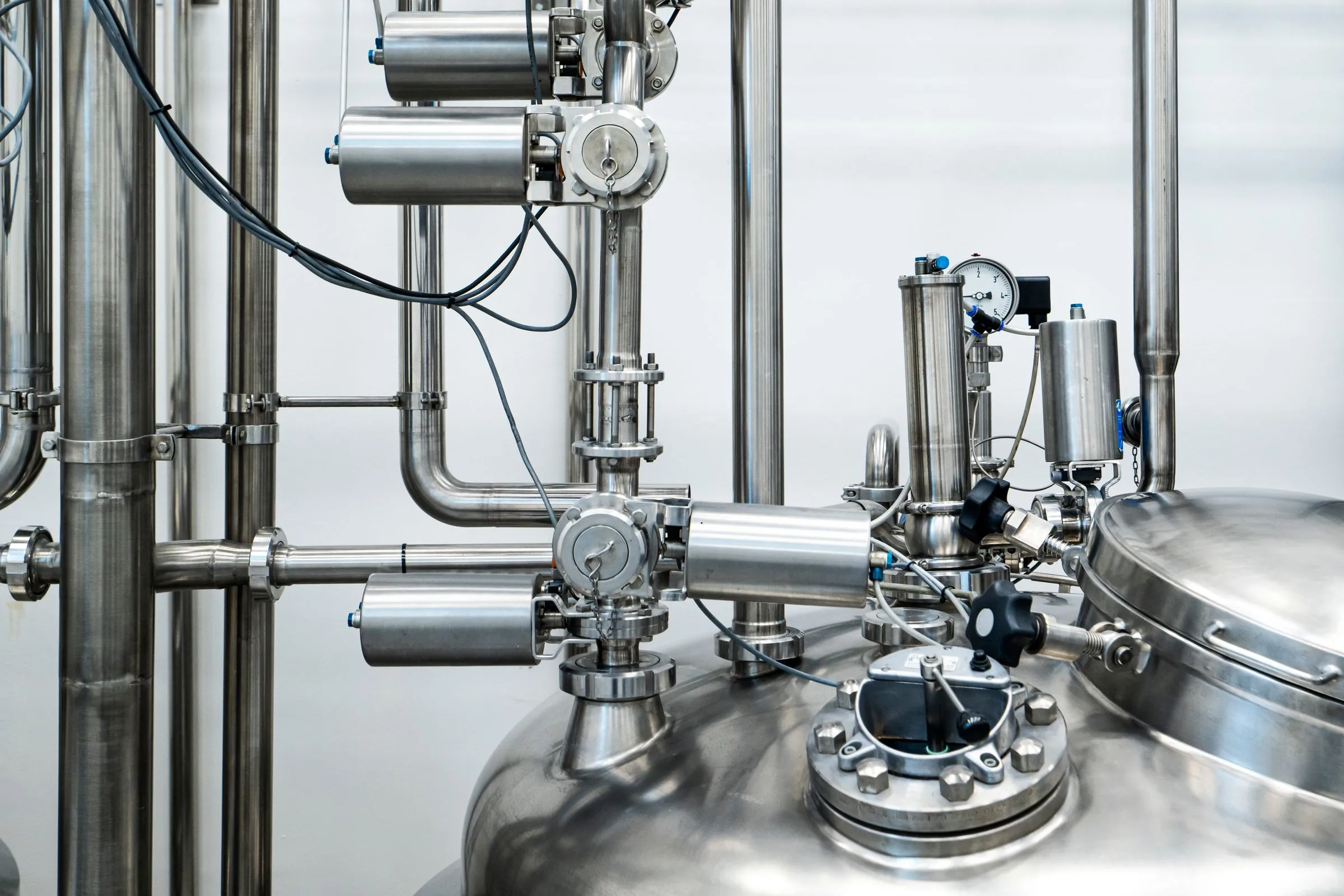 Polished stainless steel liquid-handling plant, with pipework, valves, and a pressure gauge feeding a rounded tank