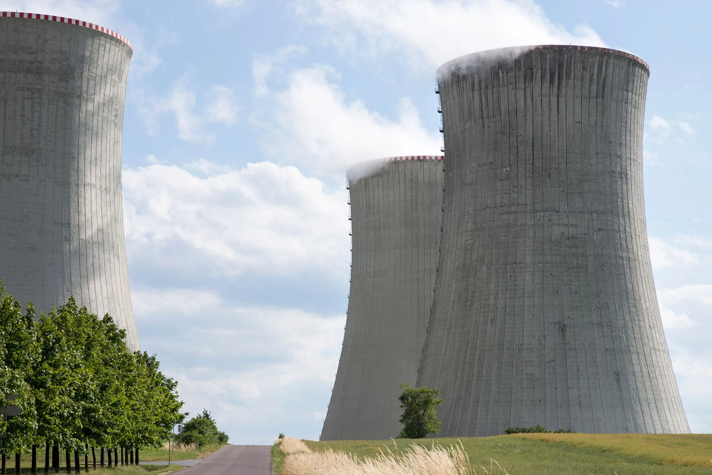 Three massive concrete hyperbolic cooling towers releasing plumes of water vapor into a partly cloudy sky above a rural road