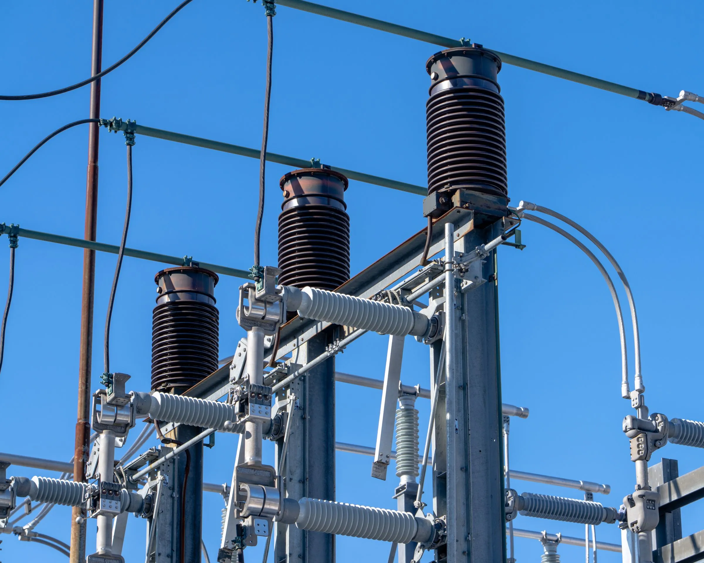 High voltage circuit breaker bushings and ribbed insulators at an electrical substation against a clear blue sky