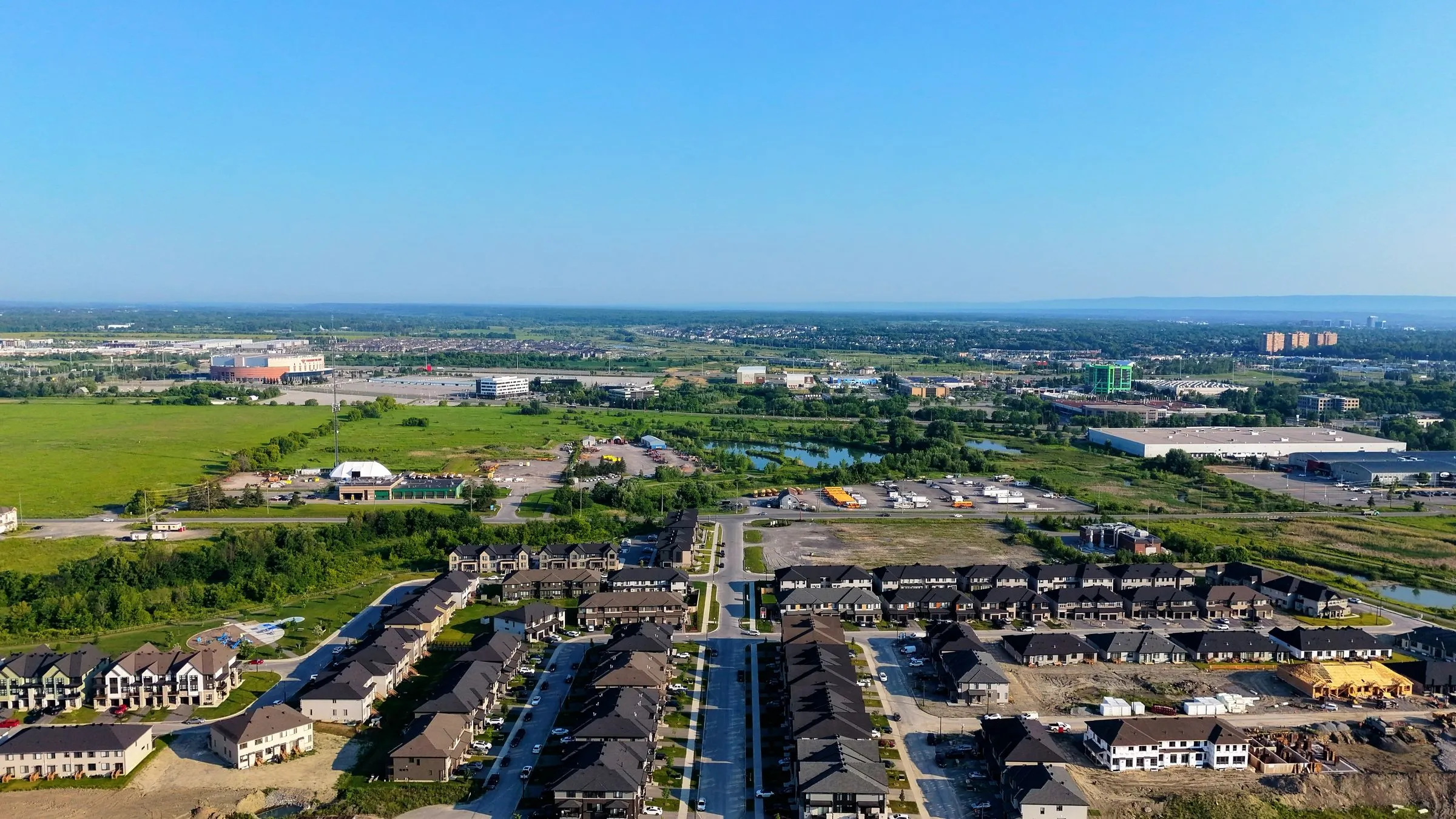 Aerial view of a new subdivision of two-story houses ending at open ground, with large flat-roofed industrial buildings on the horizon
