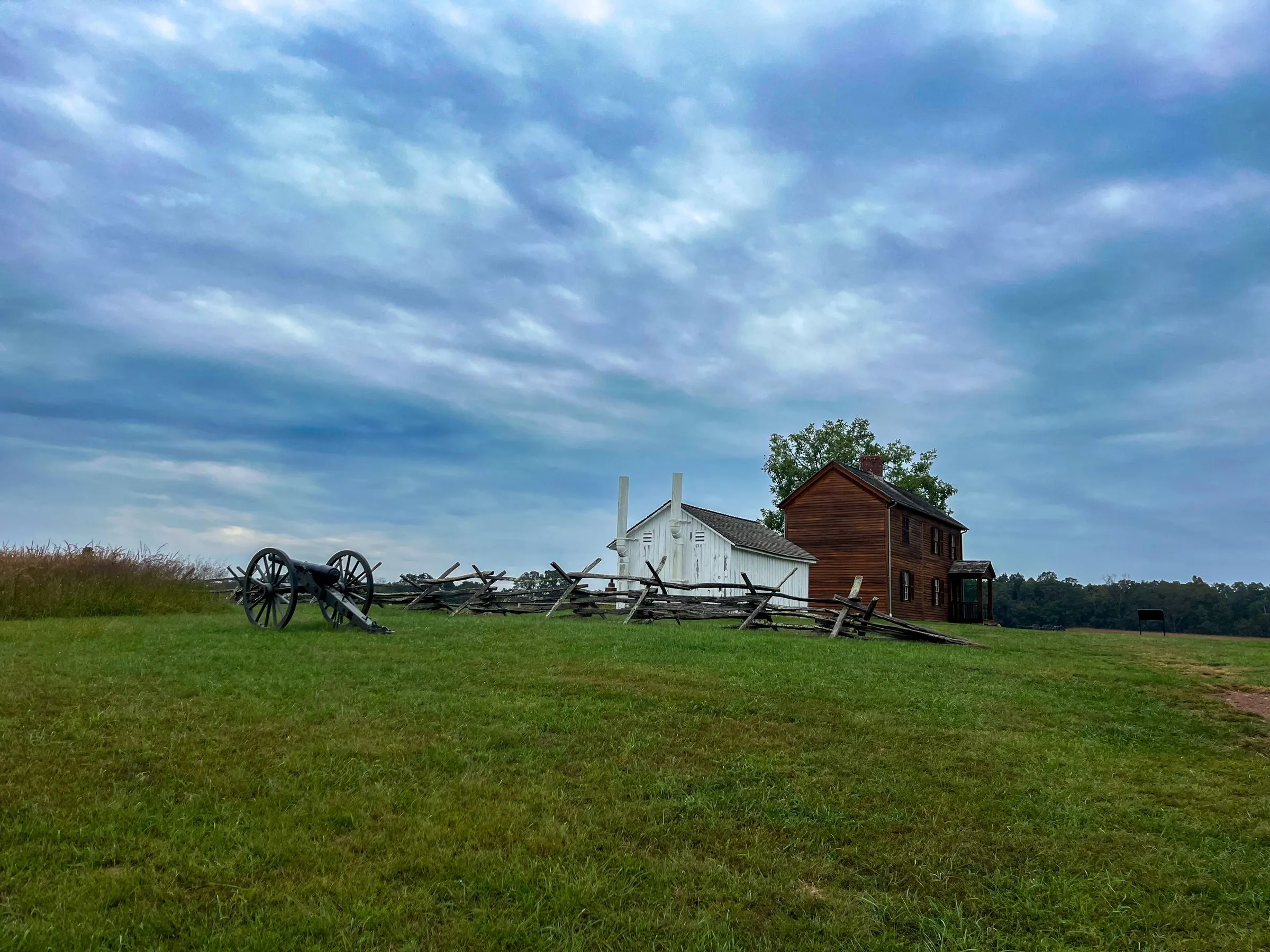 A Civil War cannon and split-rail fence in front of a restored farmhouse at Manassas National Battlefield Park in Prince William County, Virginia, under a cloudy sky