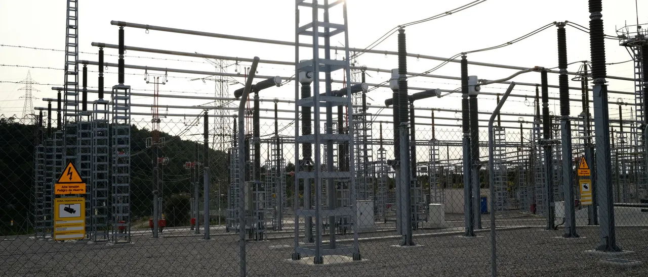 Outdoor high-voltage switchyard with steel lattice gantries, disconnect switches, and insulator strings behind a chain-link fence, the low sun flaring behind the towers