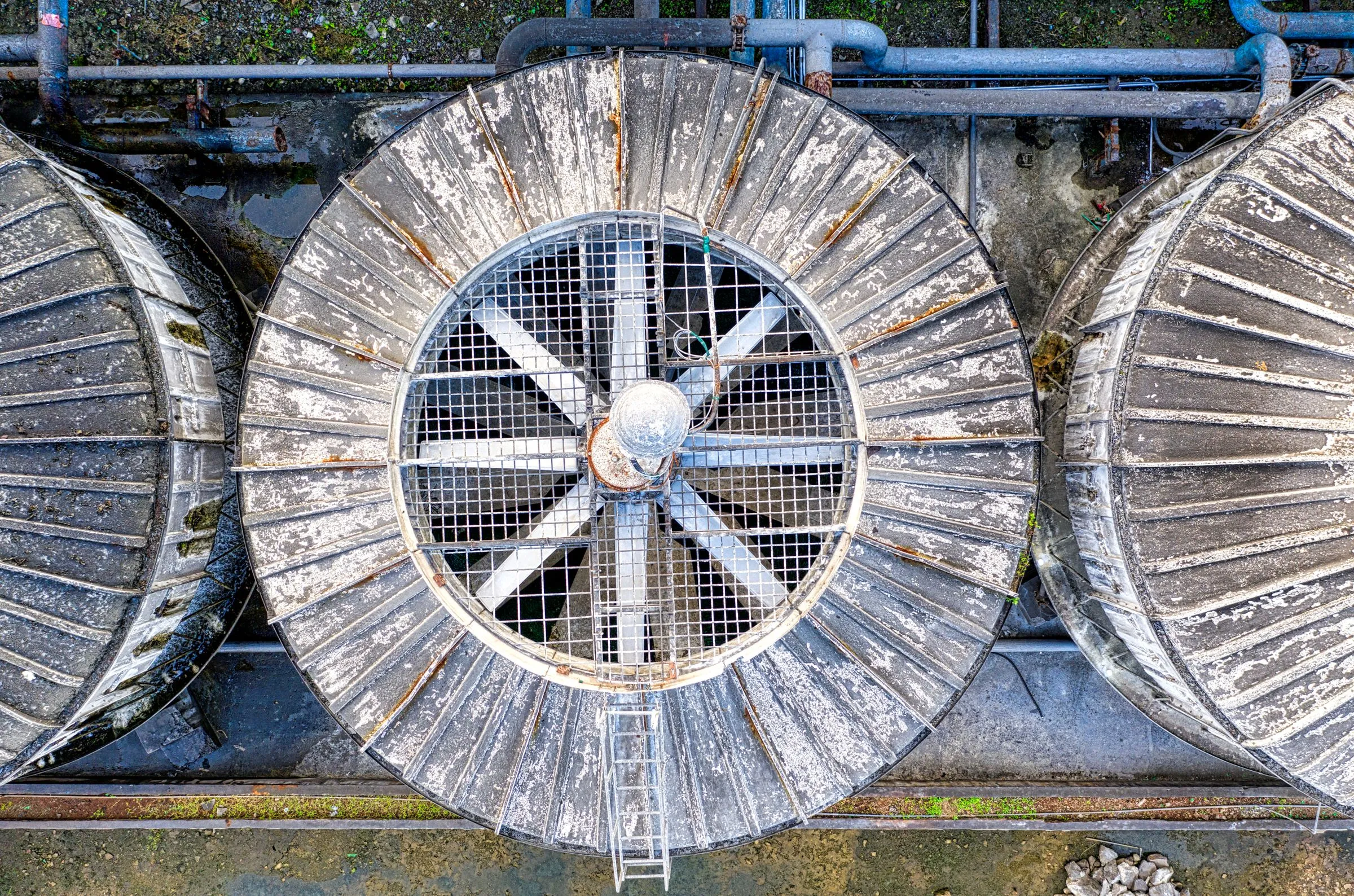 Overhead drone view of three round induced-draft cooling tower cells, one with its large fan and grating exposed, surrounded by piping