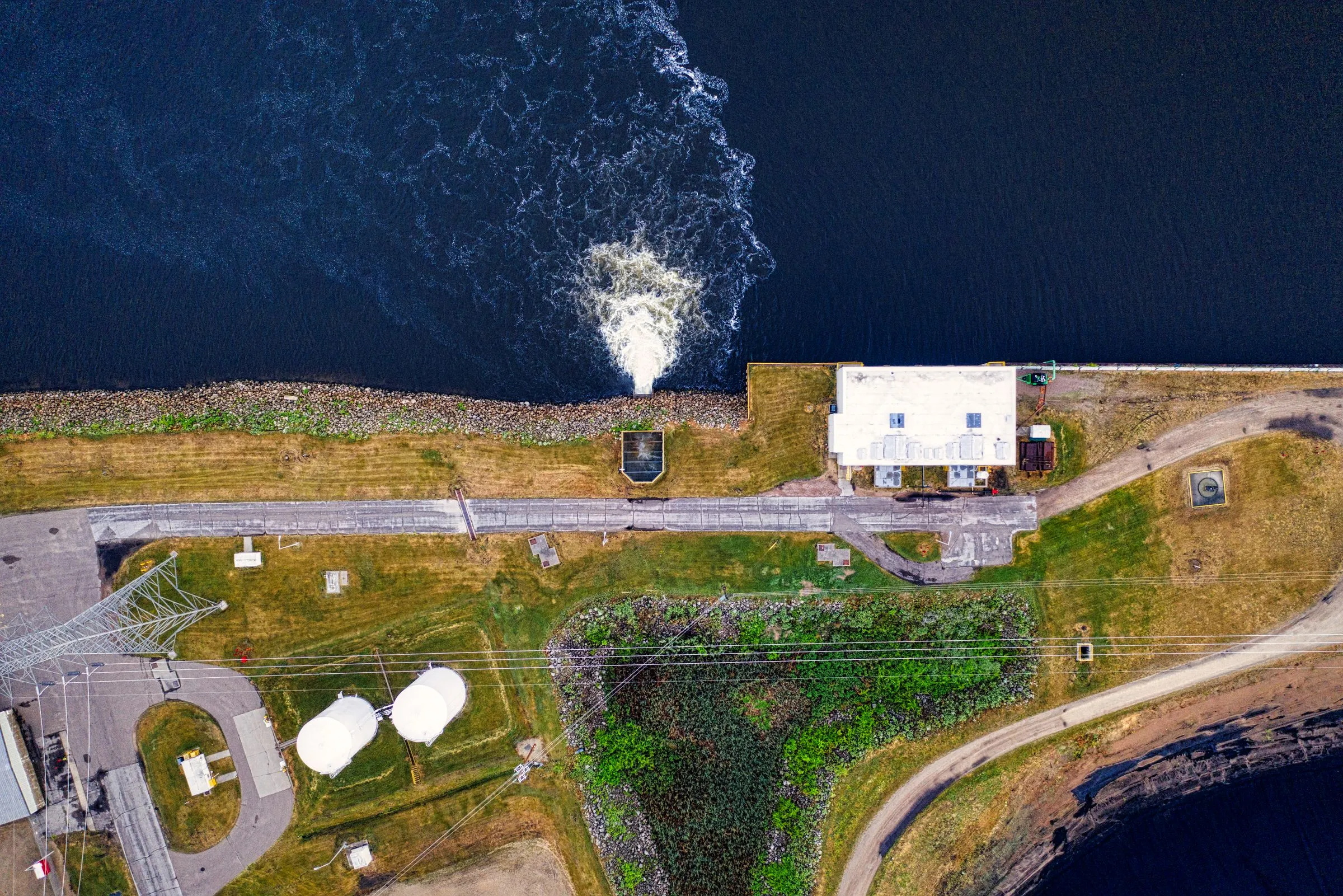 Aerial view of a power station on a riverbank, with a churning white discharge plume in dark water, storage tanks, and transmission lines