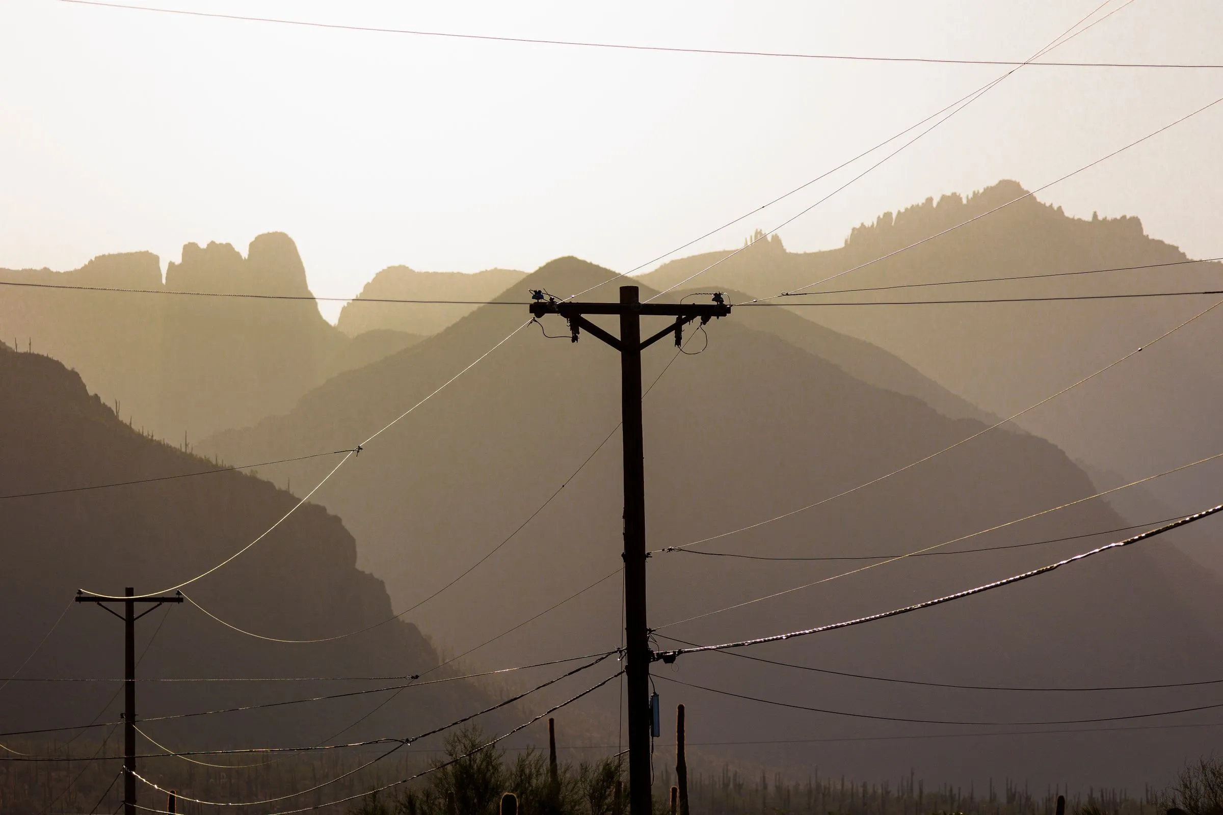 Wooden utility poles and power lines silhouetted against the hazy ridgelines of Arizona’s Superstition Mountains, with saguaro cacti below