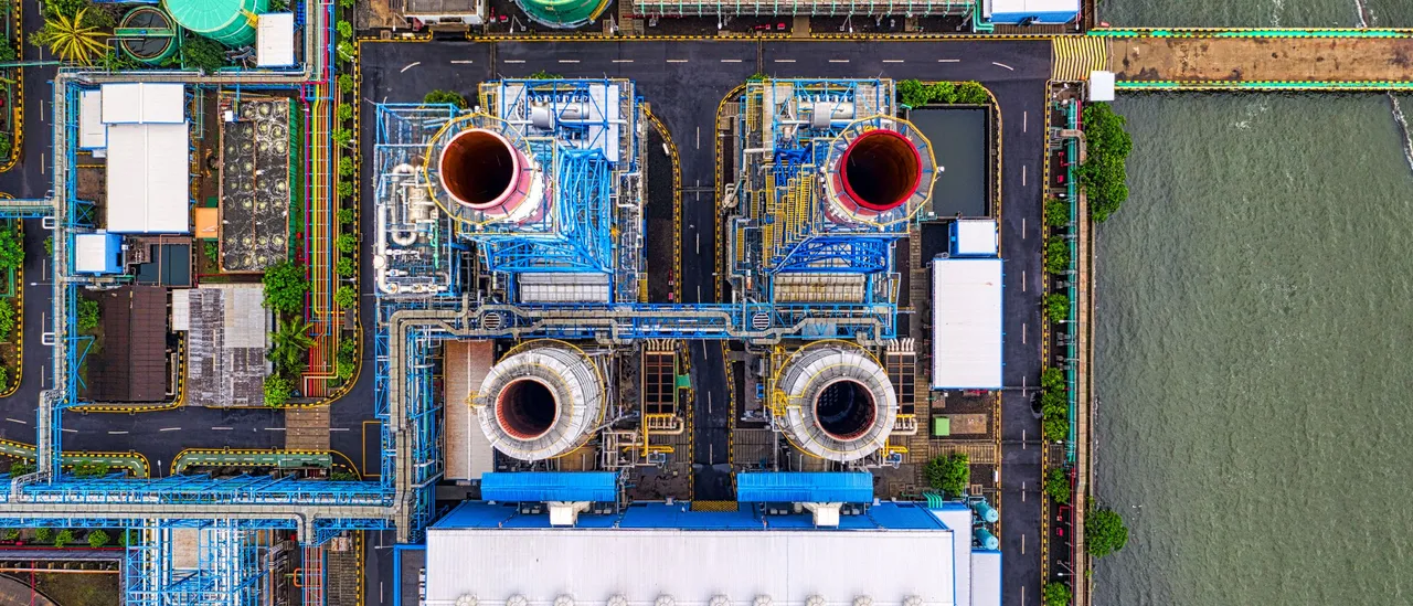 Top-down aerial of a waterfront industrial plant with a bank of cooling tower fans, storage tanks, colored piping, and four large round stacks beside the water