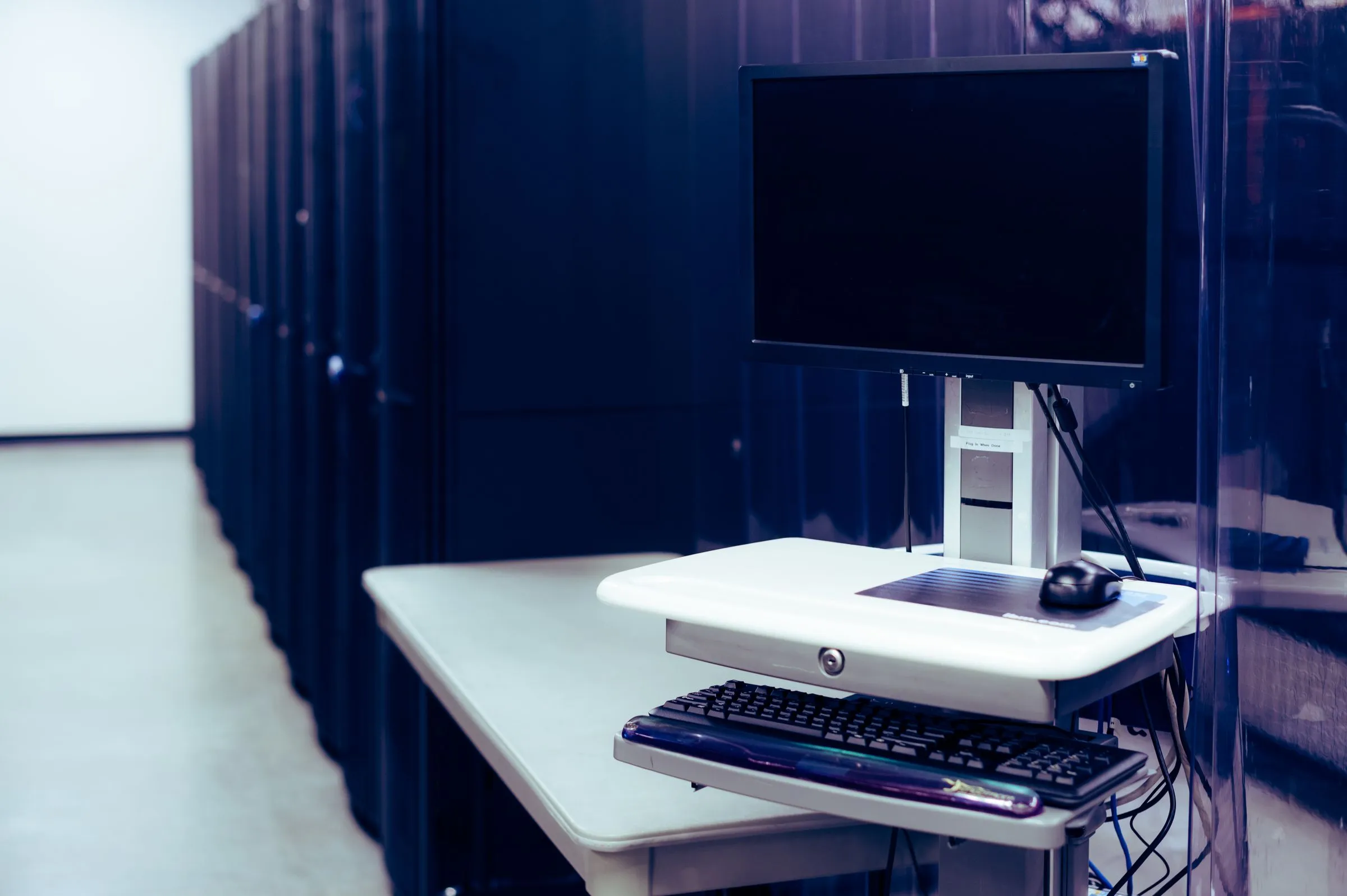 A row of dark server cabinets in a data center, with a monitor and keyboard cart seen through a transparent containment curtain