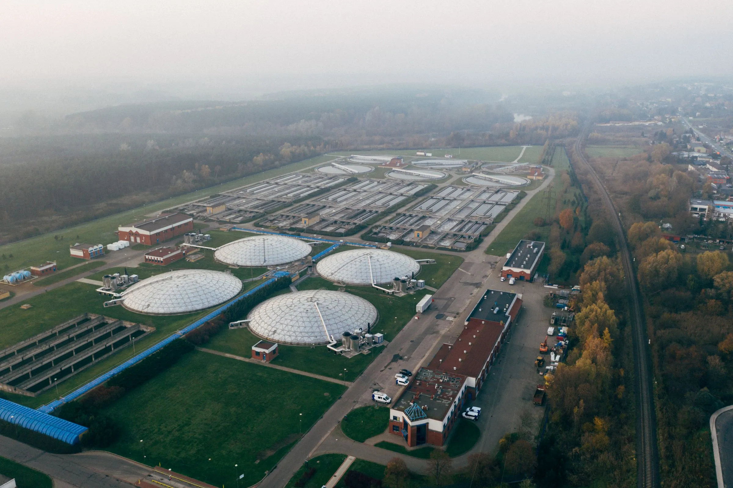 Aerial view of a water treatment plant with rows of rectangular basins, circular clarifiers, and domed digester tanks beside a rail line