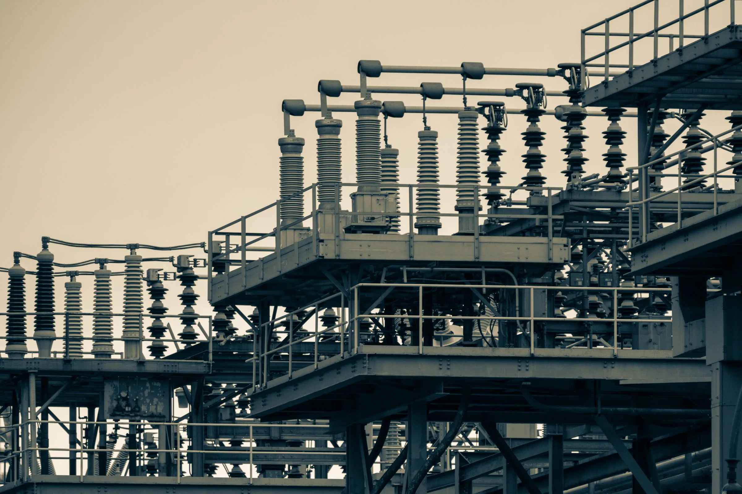 Rows of tall ribbed porcelain insulators, aluminium bus tubing, and disconnect switches mounted on steel platforms with railings at an outdoor high-voltage substation, photographed in muted sepia tones against a pale sky