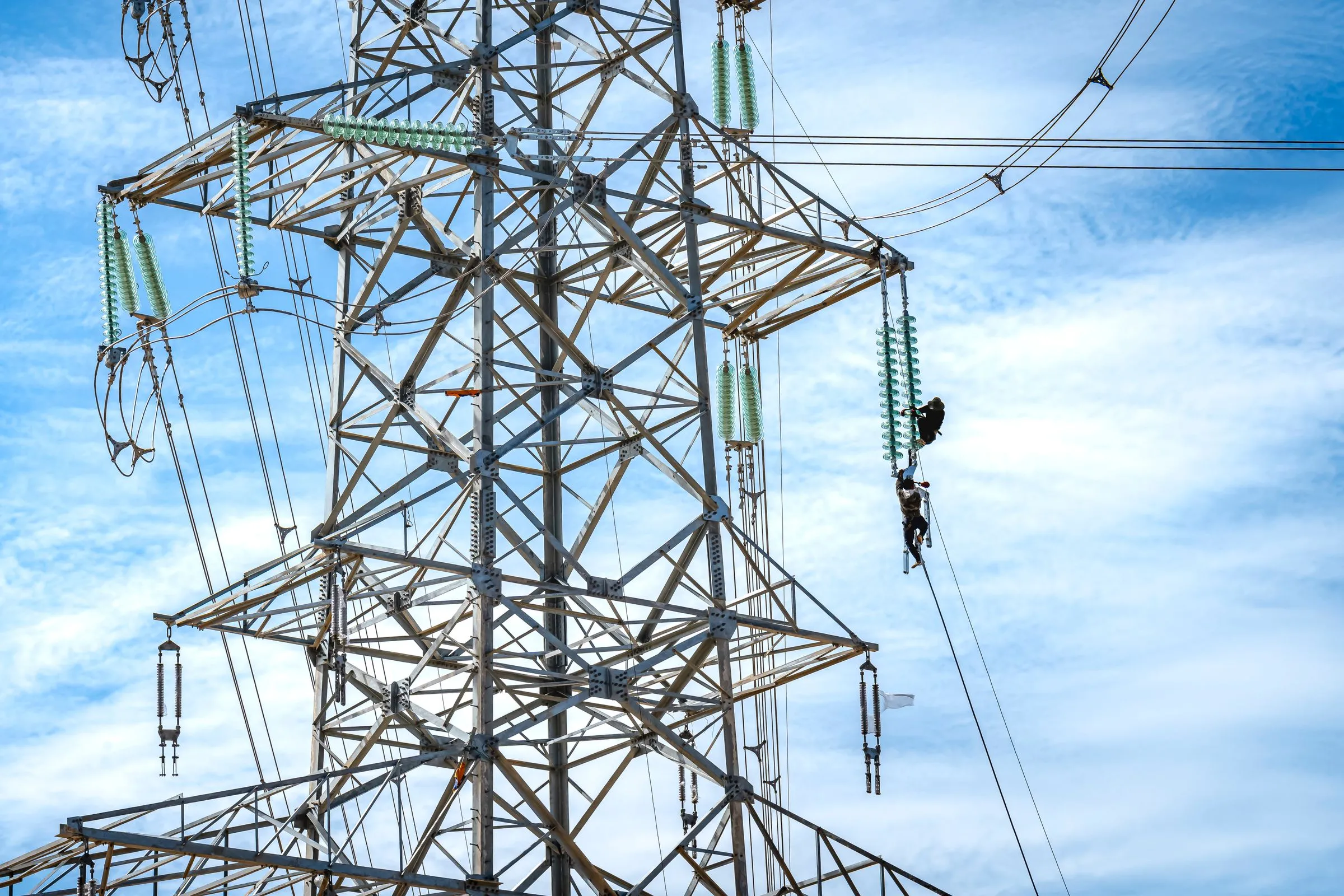 Two line workers in harnesses hanging from a string of glass insulators near the top of a tall steel lattice transmission tower, with bundled conductors and a marker flag against a blue sky streaked with cloud