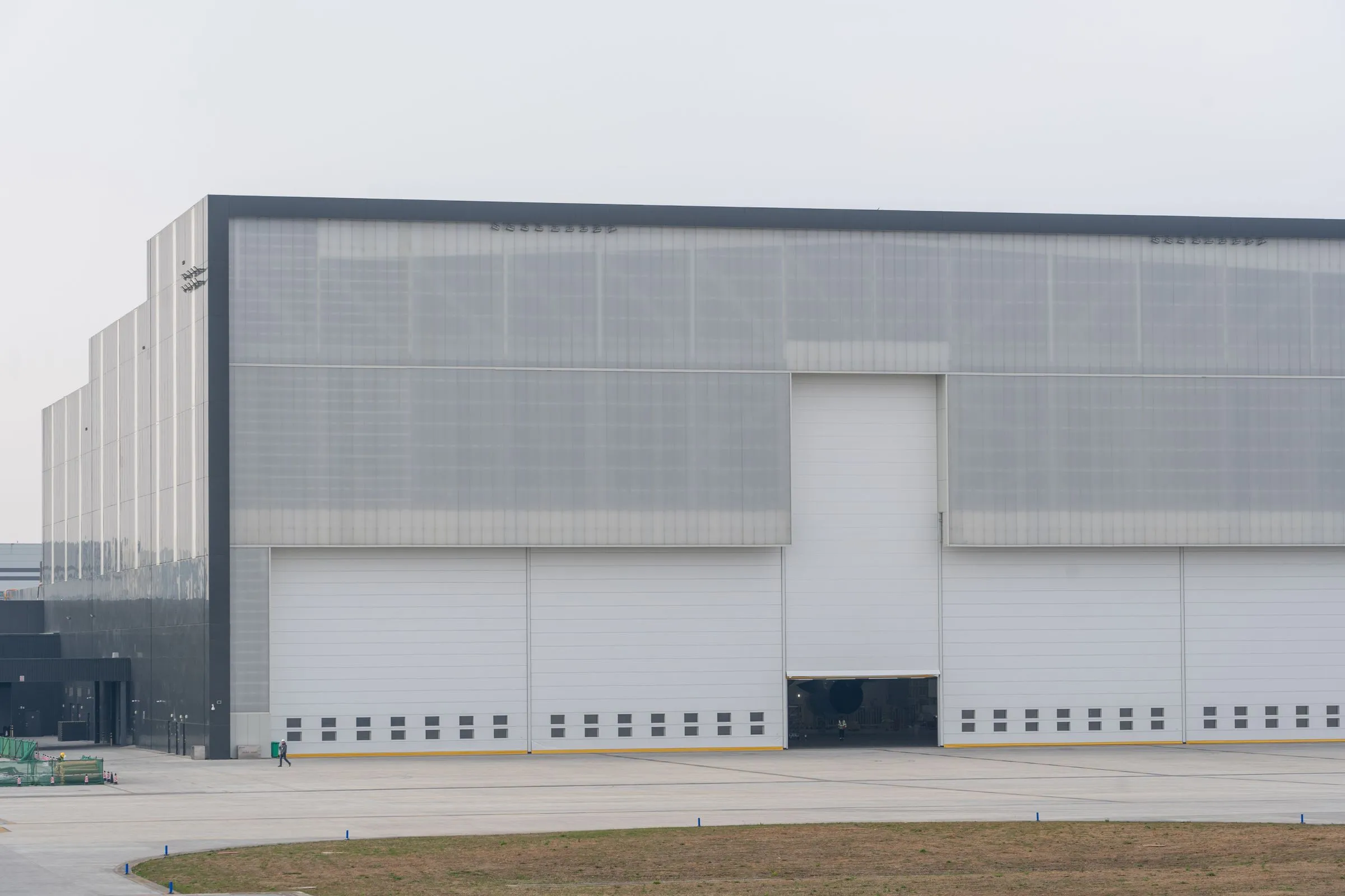 A vast windowless industrial building with a translucent gray facade and huge white doors, fronted by an empty concrete apron