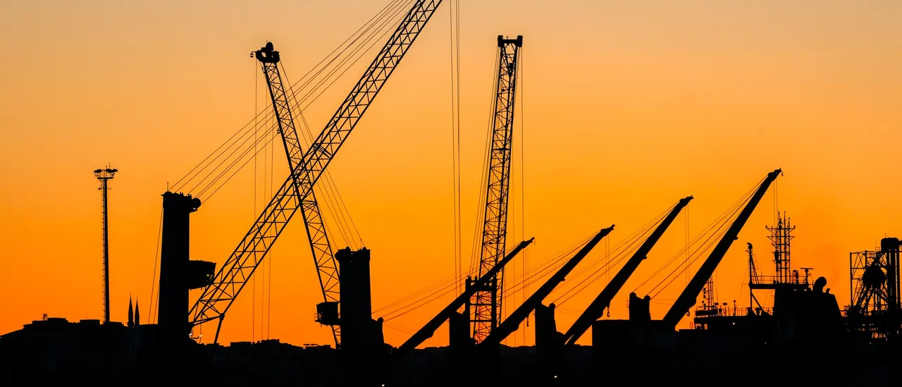 Silhouettes of tall industrial cranes against a vivid orange sunset sky