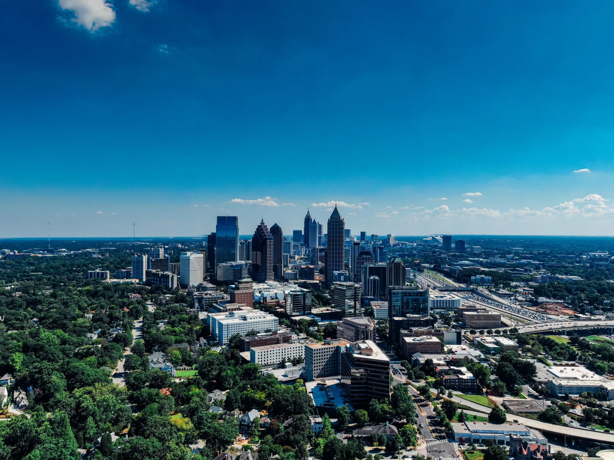 Aerial view of the Midtown Atlanta skyline under a clear blue sky, with the Downtown Connector running along the right and tree-covered neighborhoods in the foreground