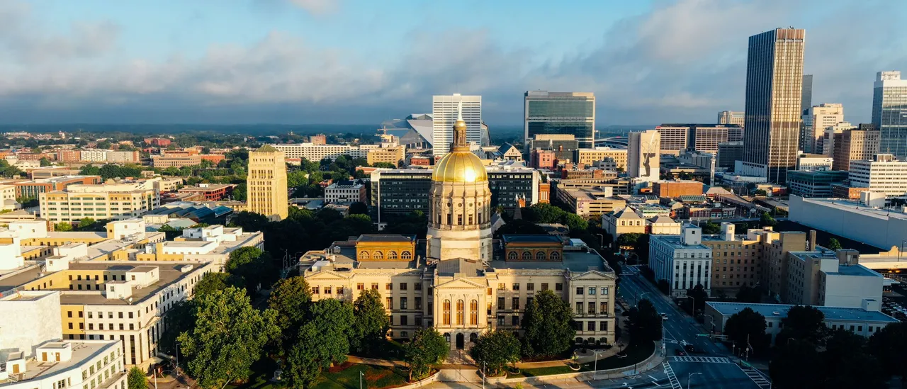 Aerial view of the Georgia State Capitol in downtown Atlanta at sunrise, its gold dome lit against a cloud bank, with the downtown skyline and Liberty Plaza behind it