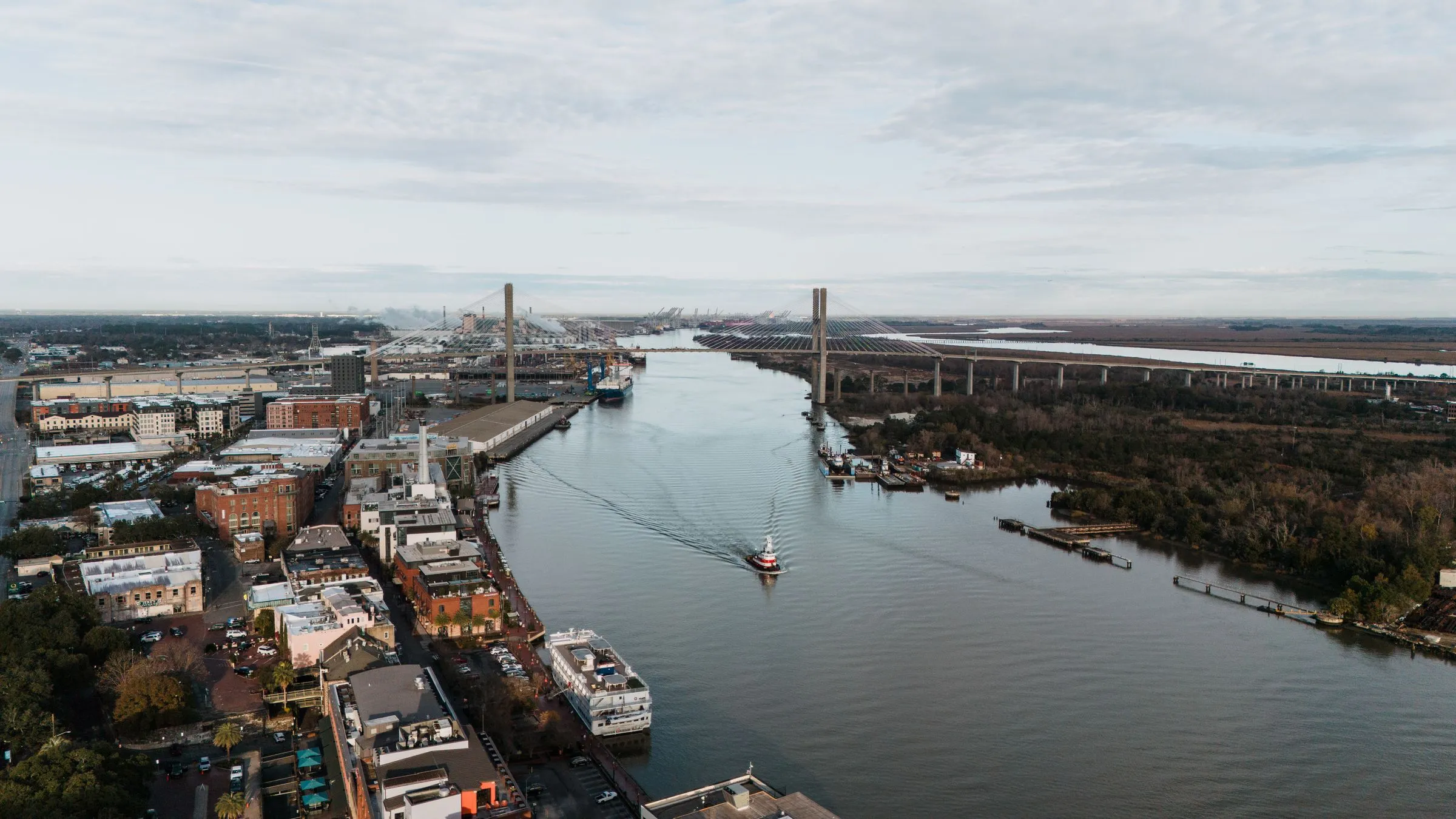 Aerial view of the Savannah River in Georgia with the cable-stayed Talmadge Memorial Bridge, a tugboat on the water, riverfront buildings on the left bank, and port cranes in the distance