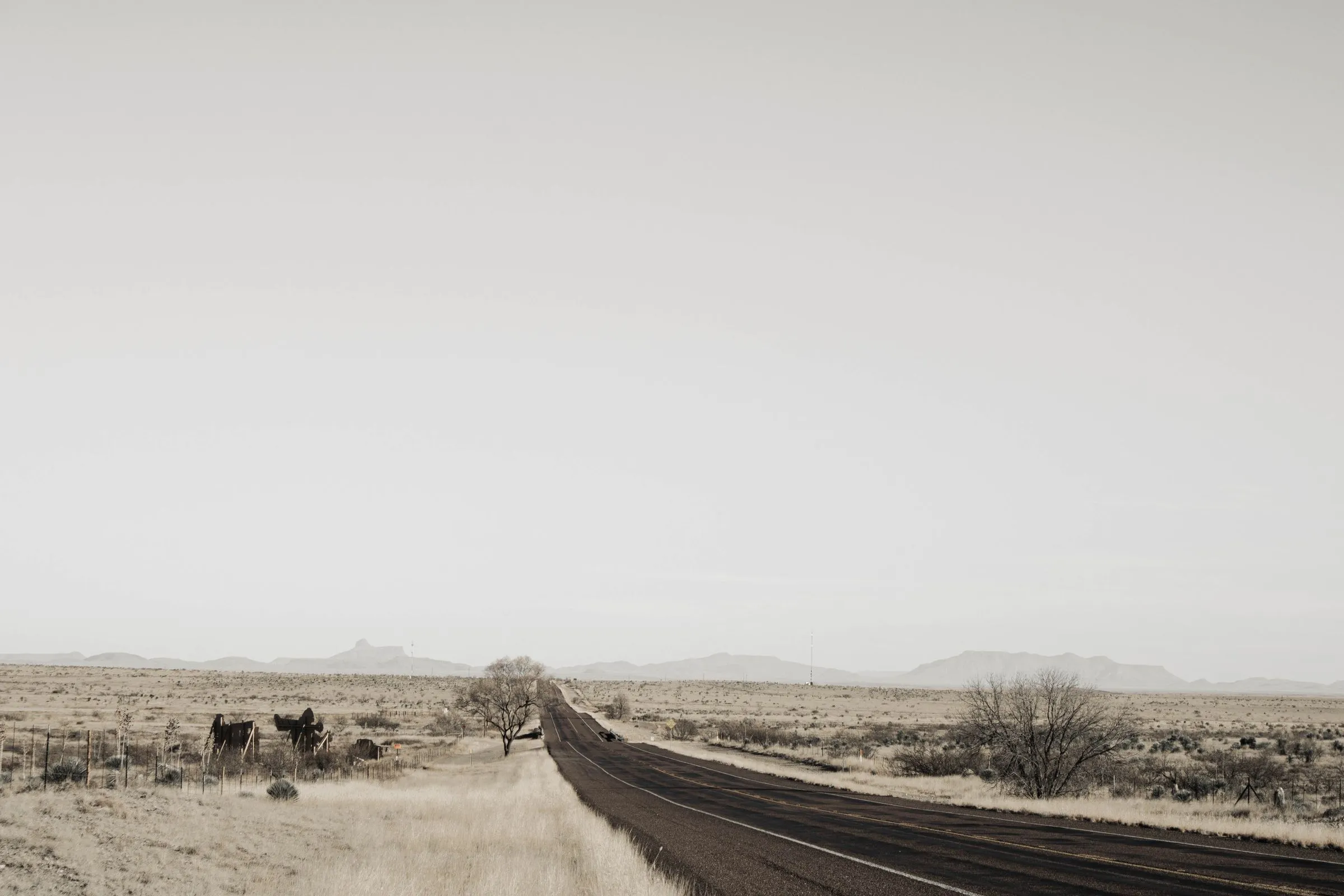 A two-lane asphalt highway running straight across flat, dry West Texas ranch land toward distant mesas under a pale sky