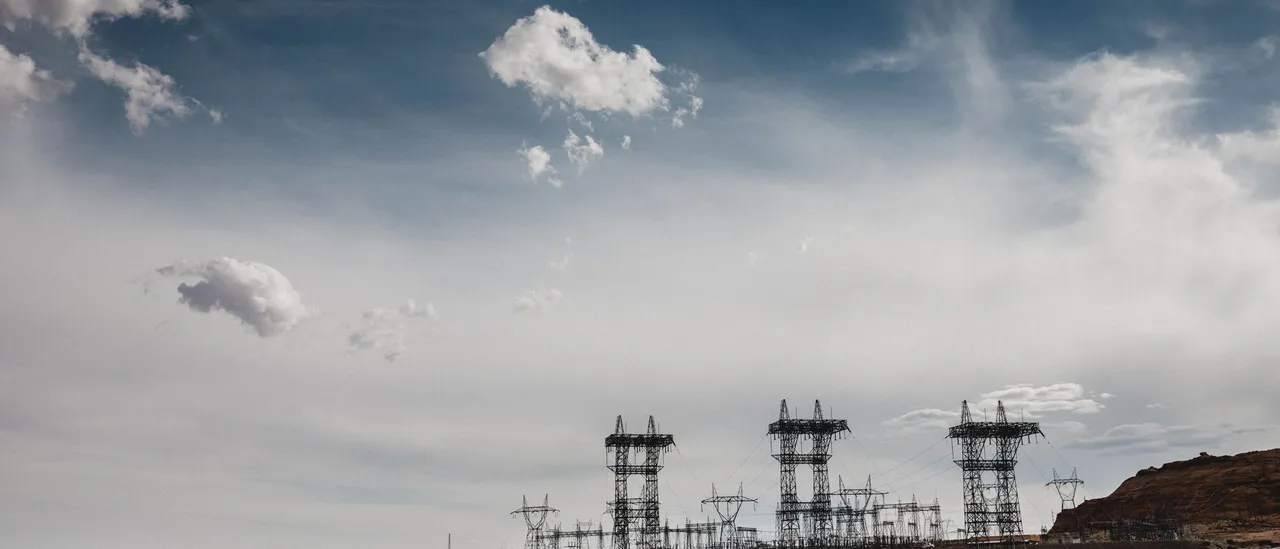 Steel high-voltage transmission towers and an electrical switchyard standing on a red-rock mesa under a partly cloudy sky in the Arizona desert