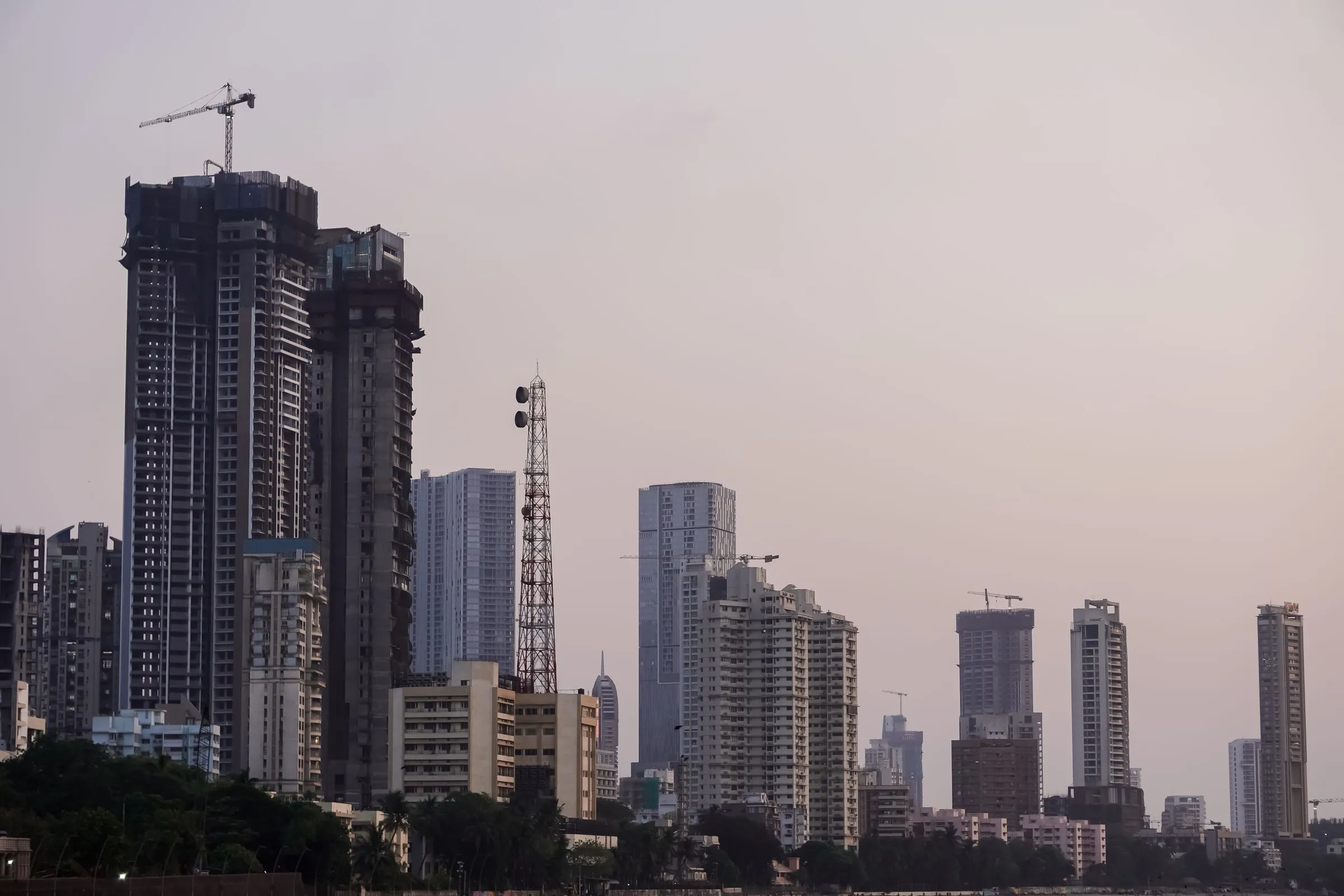 The Mumbai skyline at dusk, with high-rise towers under construction, tower cranes, and a lattice communications mast against a hazy evening sky
