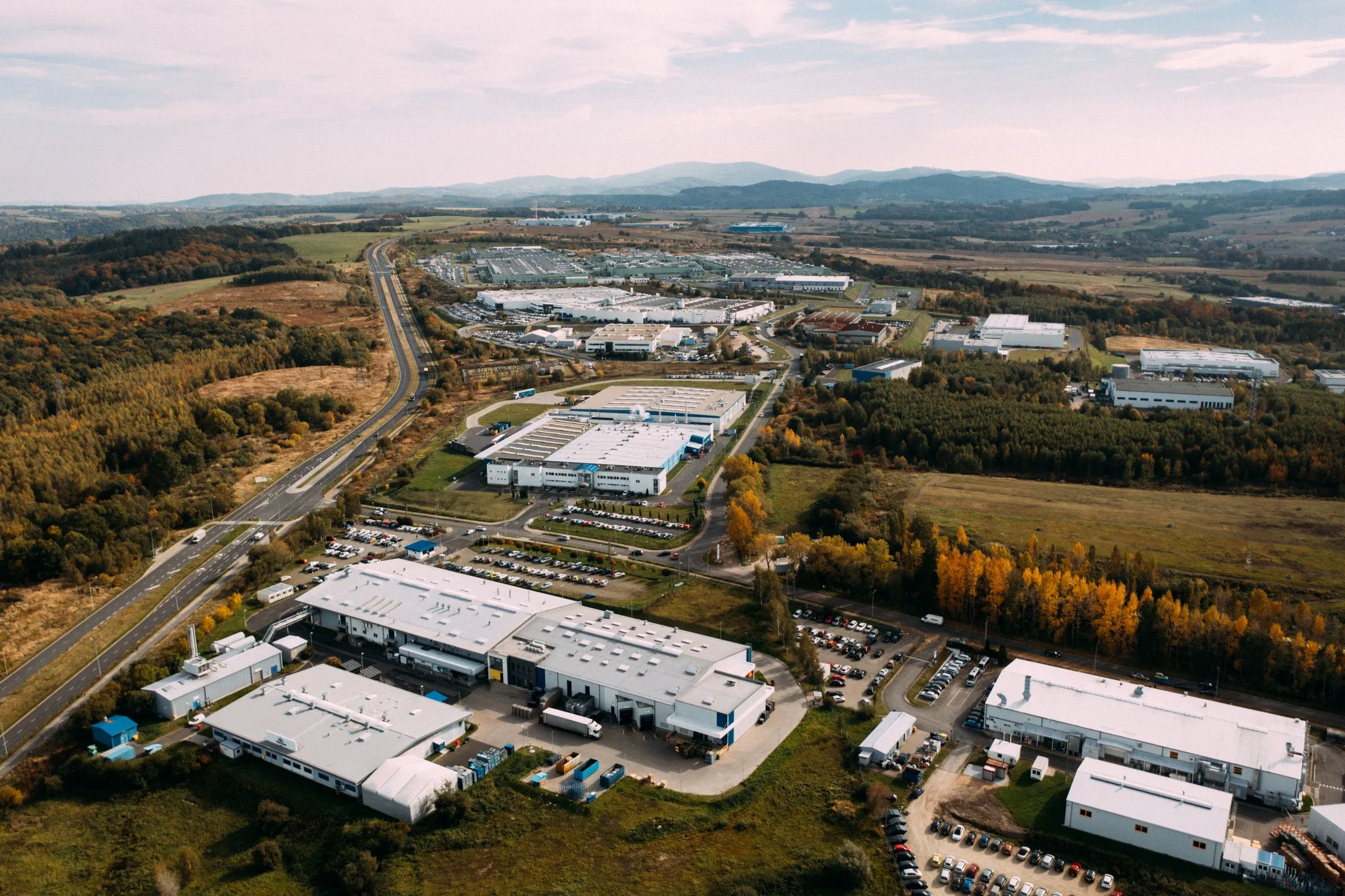 Aerial view of a sprawling industrial park, with rows of large white warehouse buildings, parking lots, and access roads spreading across open countryside toward distant hills
