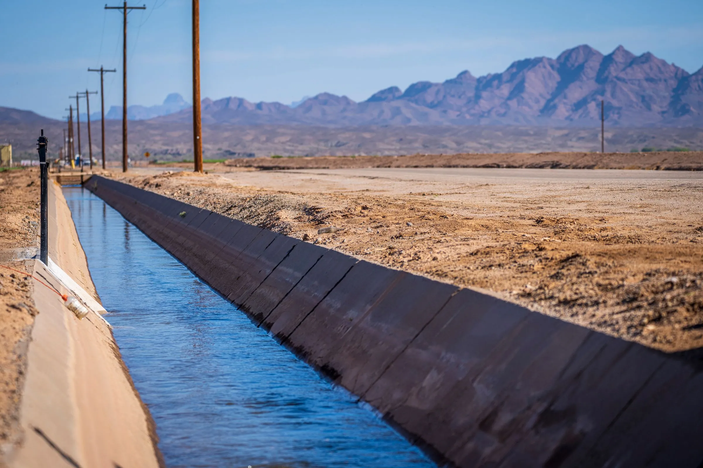 A concrete-lined irrigation canal full of blue Colorado River water running straight through bare desert farmland toward jagged purple mountains near Yuma, Arizona, with wooden power poles along the bank