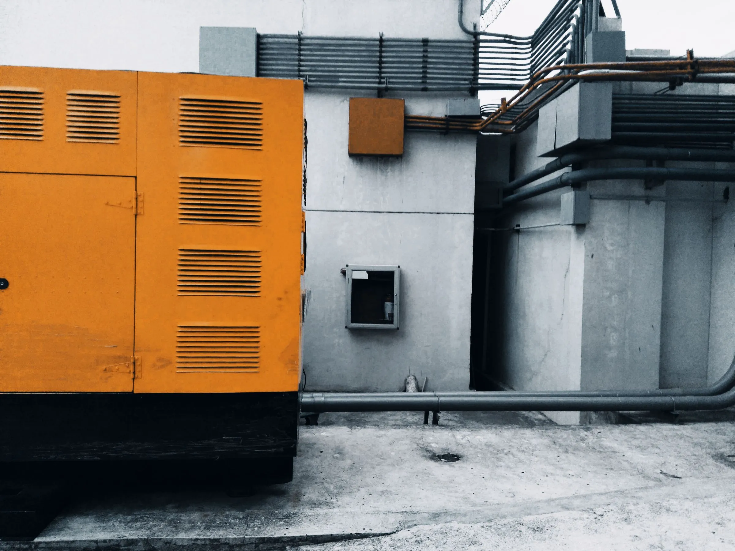 A yellow steel generator enclosure with louvered cooling vents standing on a concrete pad against a grey industrial wall, with exhaust and conduit piping running overhead