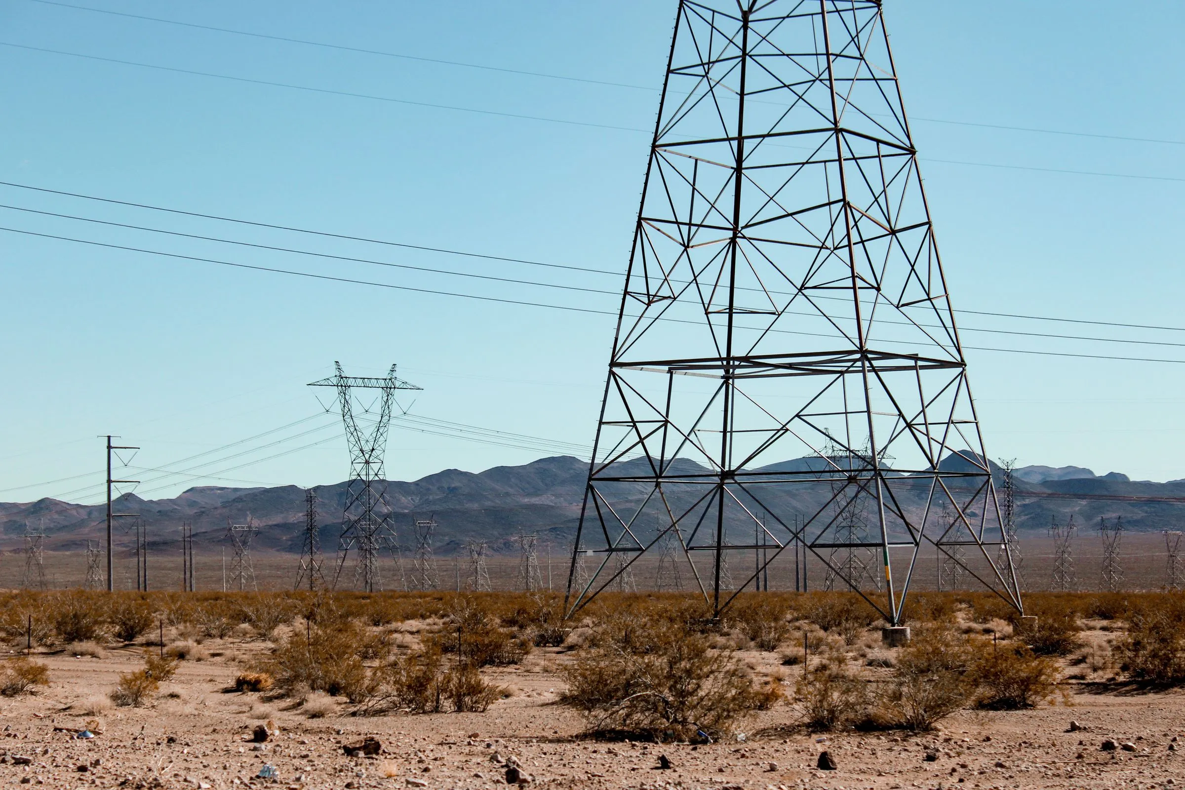 Steel lattice transmission towers carrying high-voltage lines across a flat scrub-covered Nevada desert valley, with a range of bare brown mountains under a clear blue sky