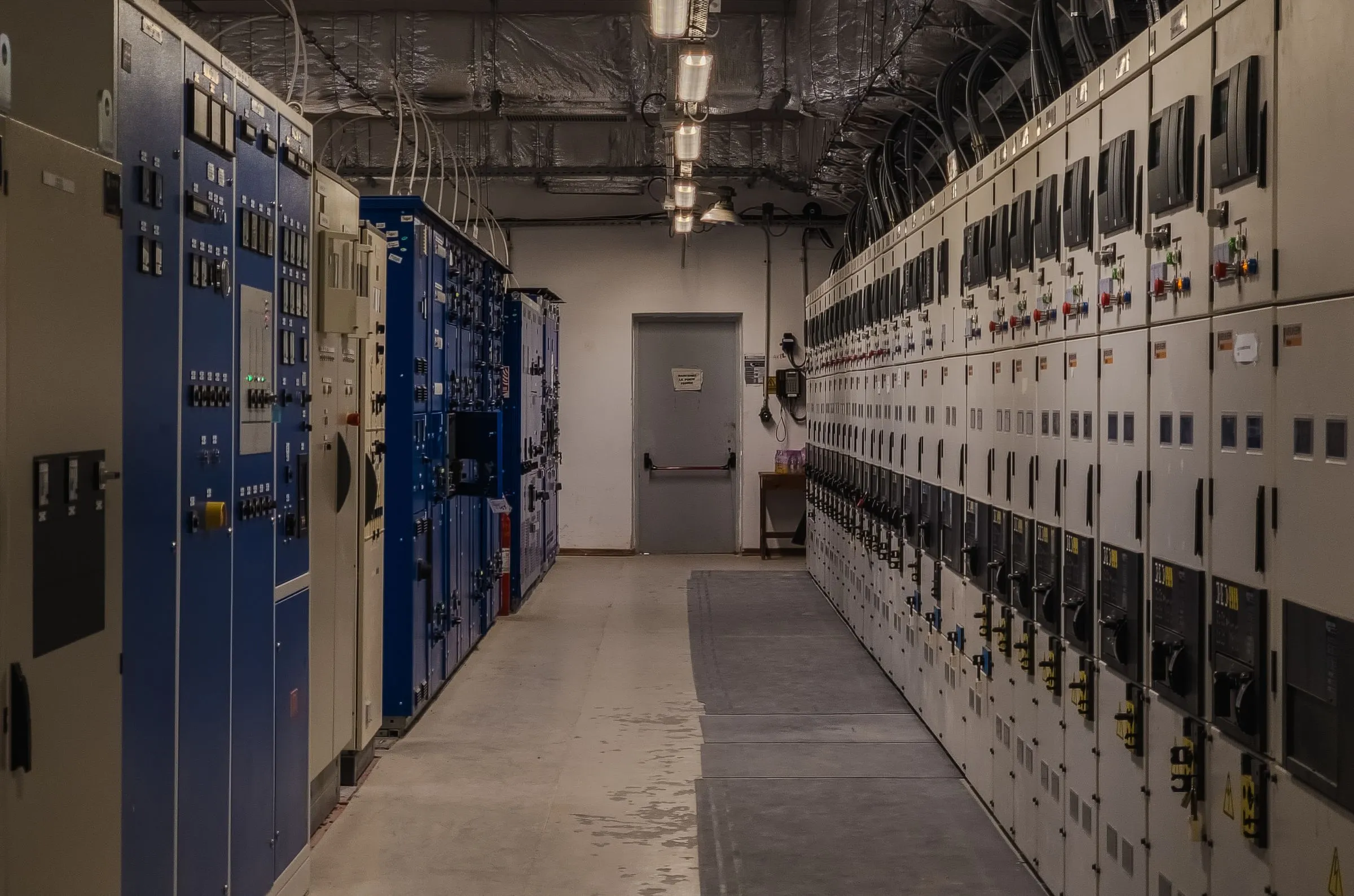 Rows of electrical switchgear cabinets lining both sides of a narrow aisle in an industrial power distribution room