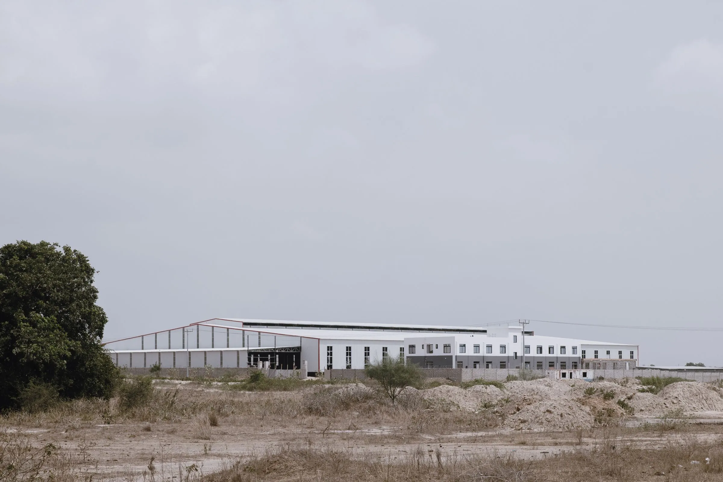 A long, low white industrial building rising from a dry rural landscape, with mounds of excavated dirt in the foreground