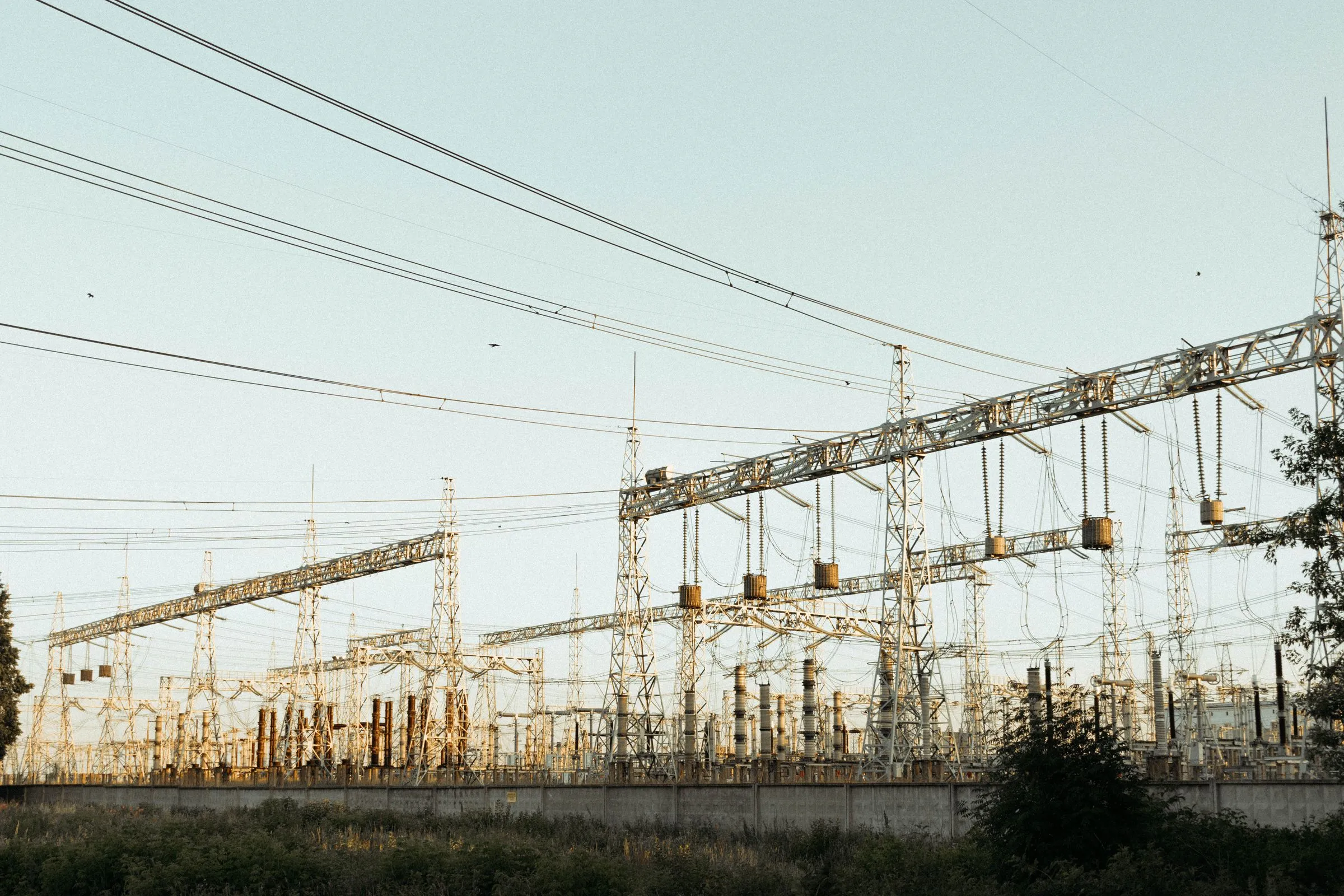 A high-voltage electrical substation with lattice steel towers, transformers, and overhead lines in evening light