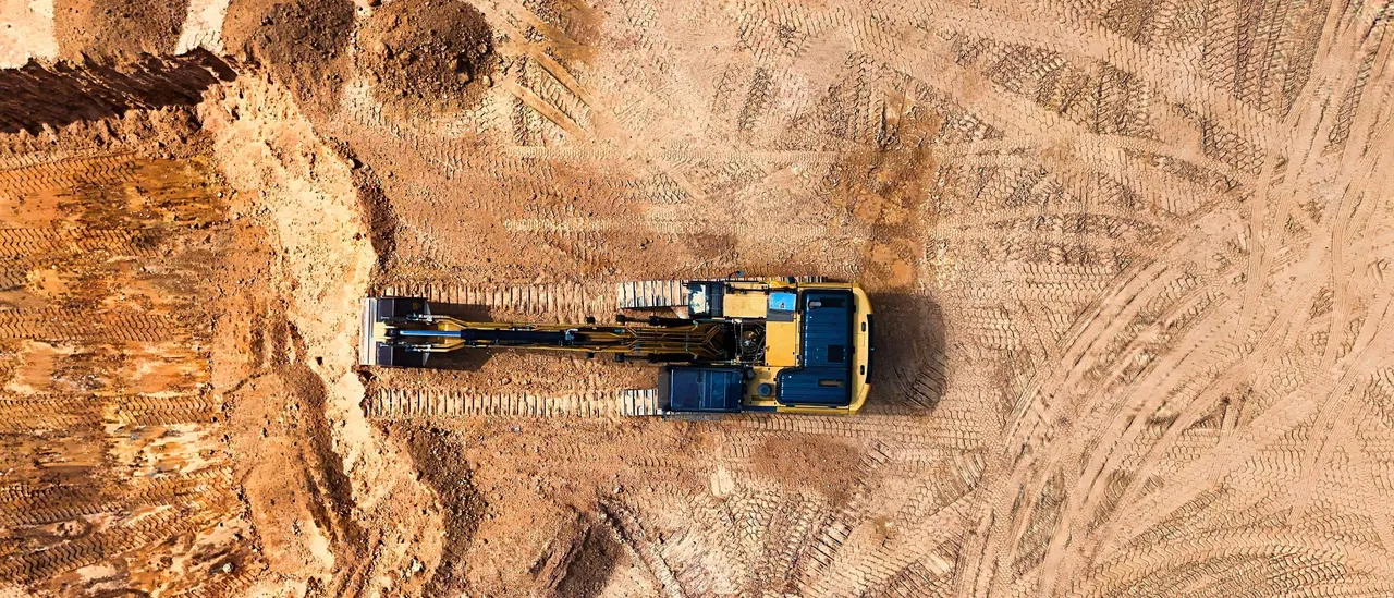 Overhead view of a yellow excavator working on a large graded dirt construction site covered in tire tracks