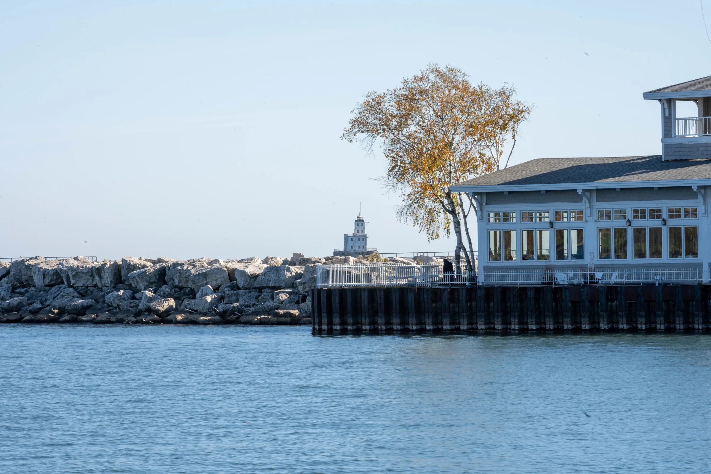 The Milwaukee lakefront on a clear autumn day, with a limestone breakwater, the white Milwaukee Breakwater Lighthouse on the horizon, a yellow-leaved birch, and a white clapboard harbor pavilion with a gray shingled roof standing on steel pilings over the blue water of Lake Michigan